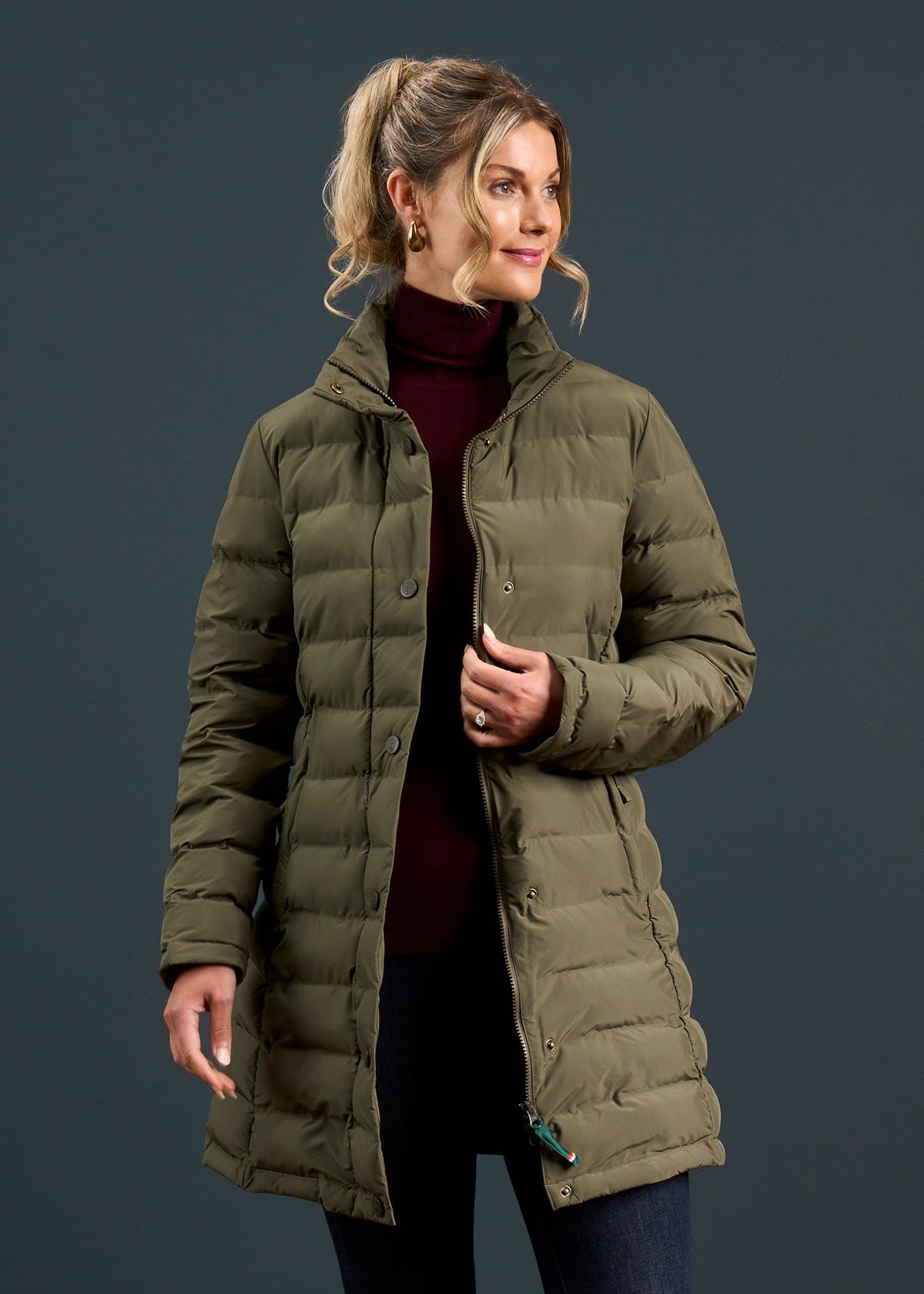 Woman in a Calsall Ladies Coat in Olive, dark jeans, and maroon turtleneck stands against a dark background.