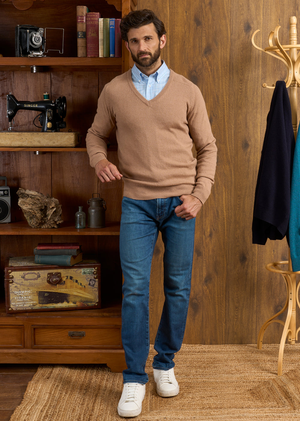 A man in the Hampshire Lambswool Vee Neck Jumper in Camel stands by wooden shelves in a cozy room.