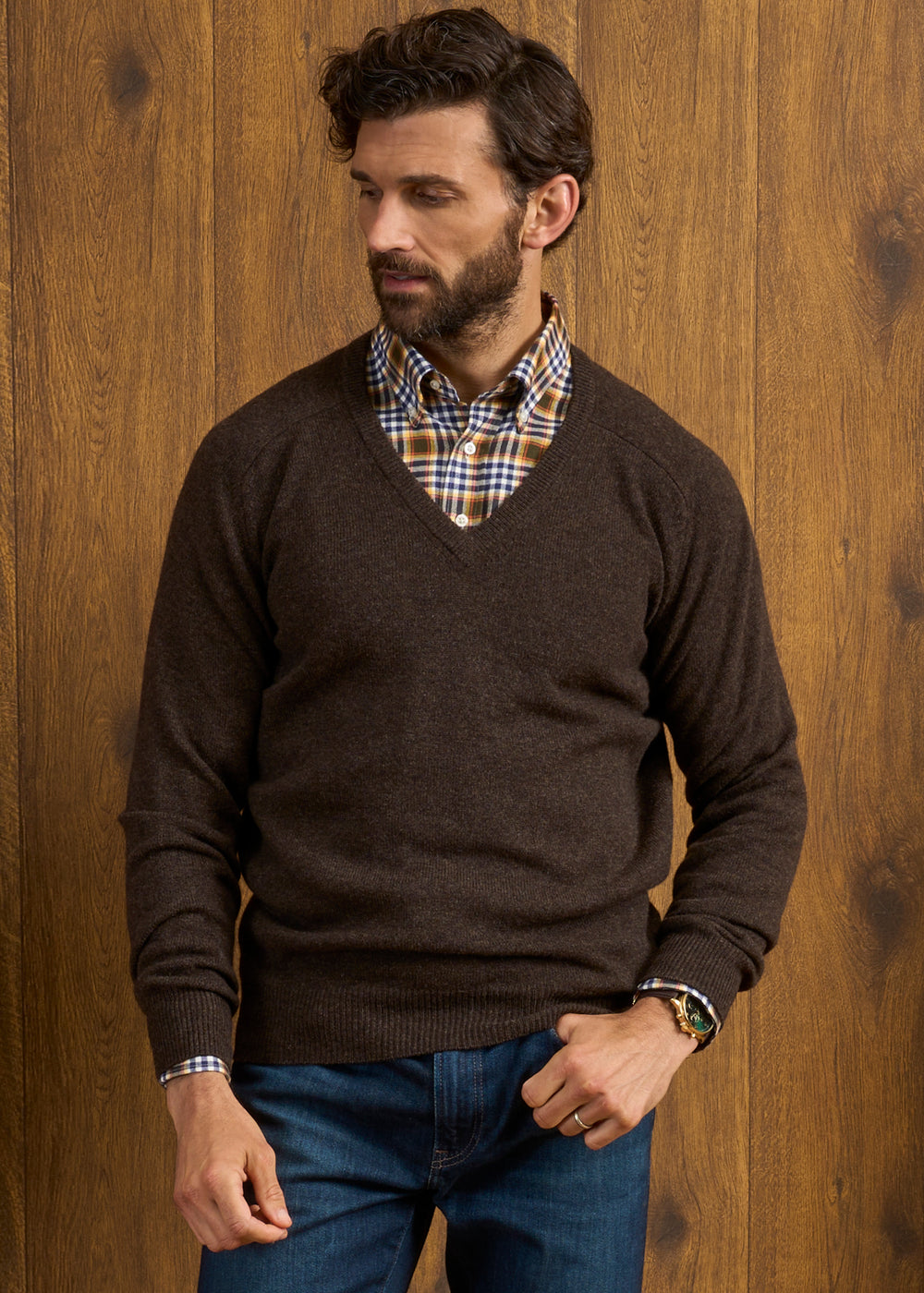 Man wears Hampshire Lambswool Vee Neck Jumper in Cocoa over a plaid shirt, standing by a wooden wall looking sideways.