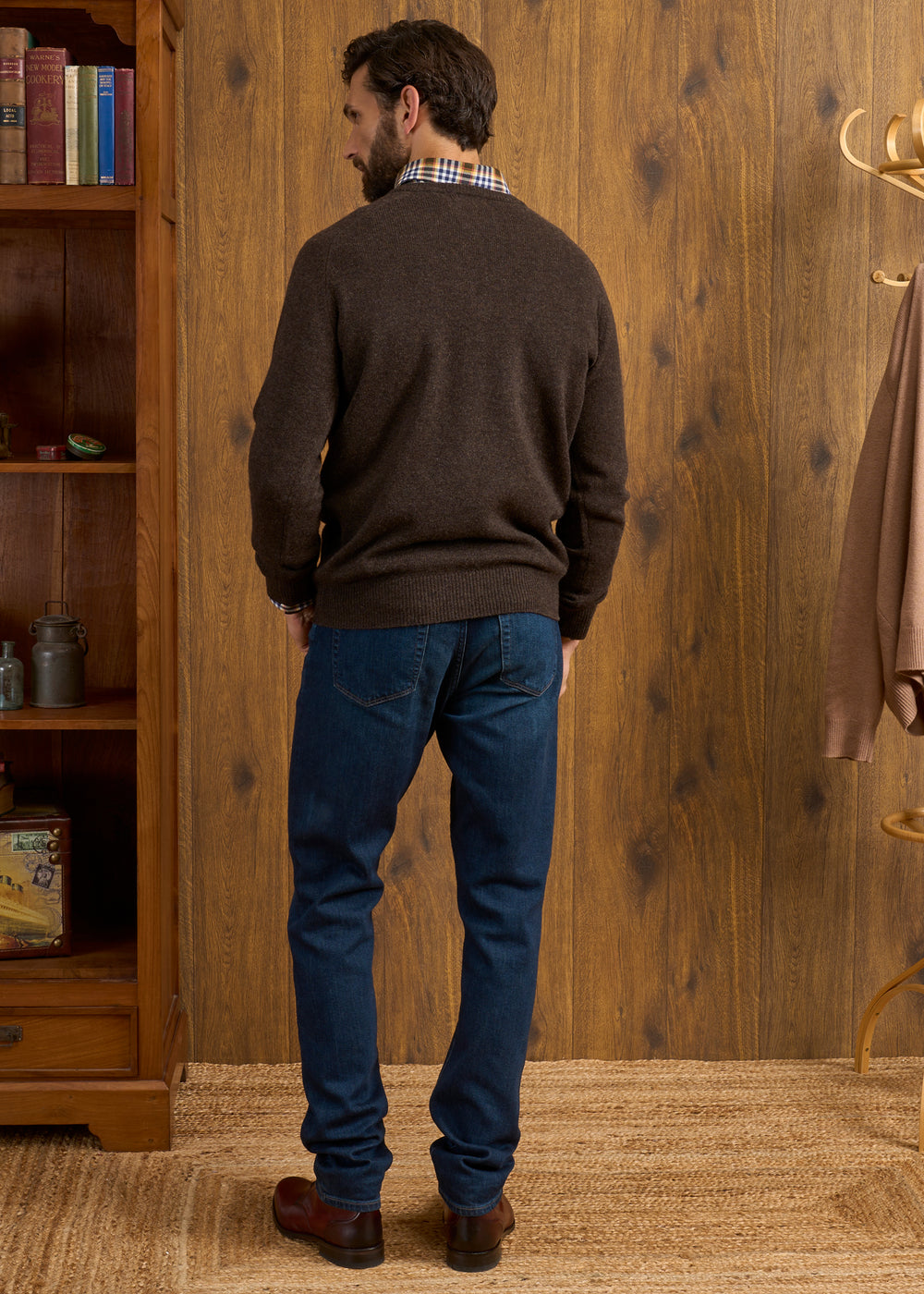 Man indoors with back turned, wearing Hampshire Lambswool Vee Neck Jumper in Cocoa and blue jeans on woven rug.