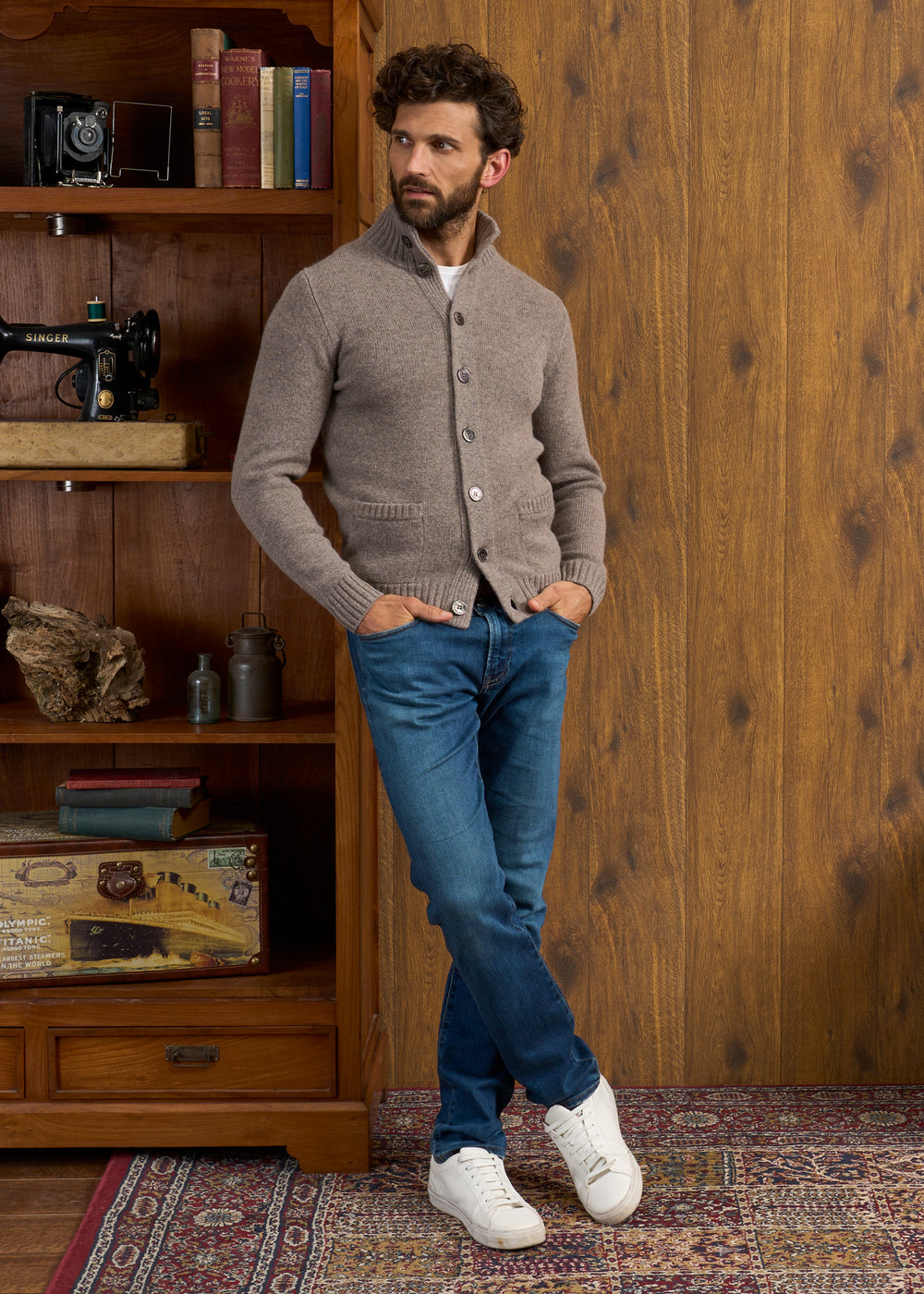 Man wears Landford Lambswool Buttoned Mock Neck Jumper in Vole, blue jeans, by bookshelf on patterned rug.