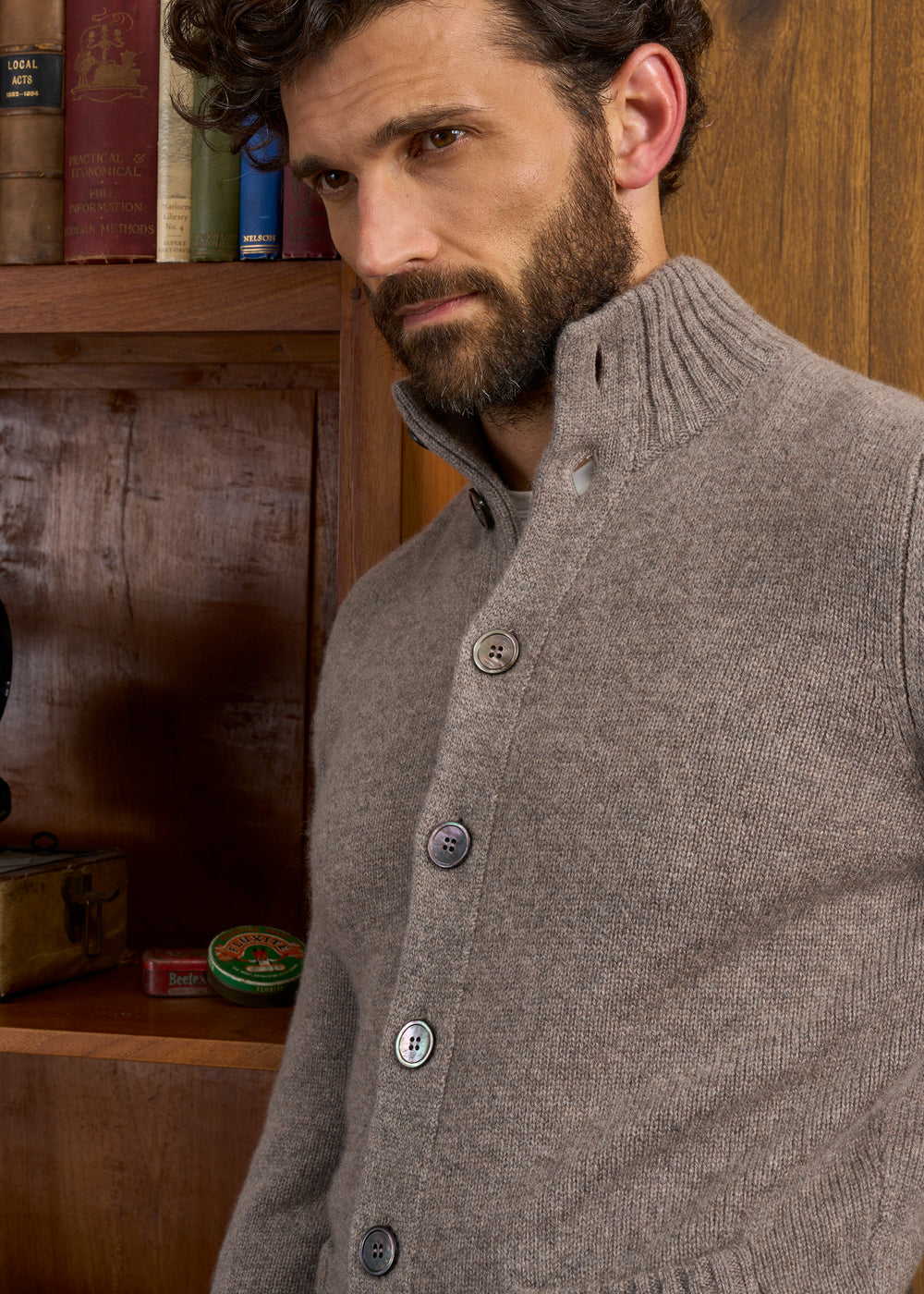 Man wears Landford Lambswool Buttoned Mock Neck Jumper in Vole, standing by a shelf with books and vintage items.