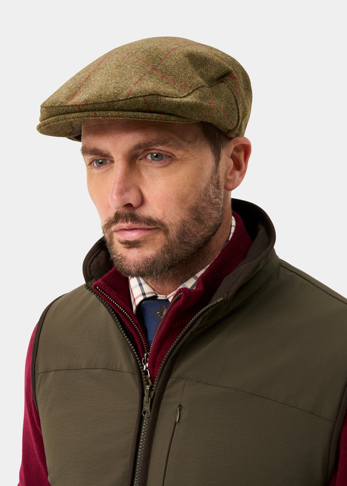 Man wears Combrook Men’s Tweed Waterproof Flat Cap in Sage with olive vest, burgundy sweater, and plaid shirt.