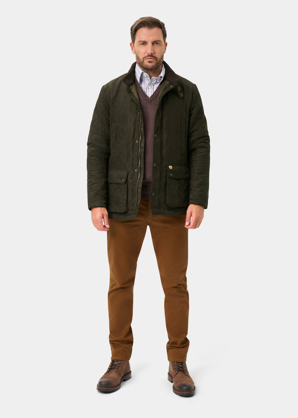 Man wearing Felwell Men’s Quilted Jacket in Olive, brown pants, boots, and v-neck sweater over collared shirt.