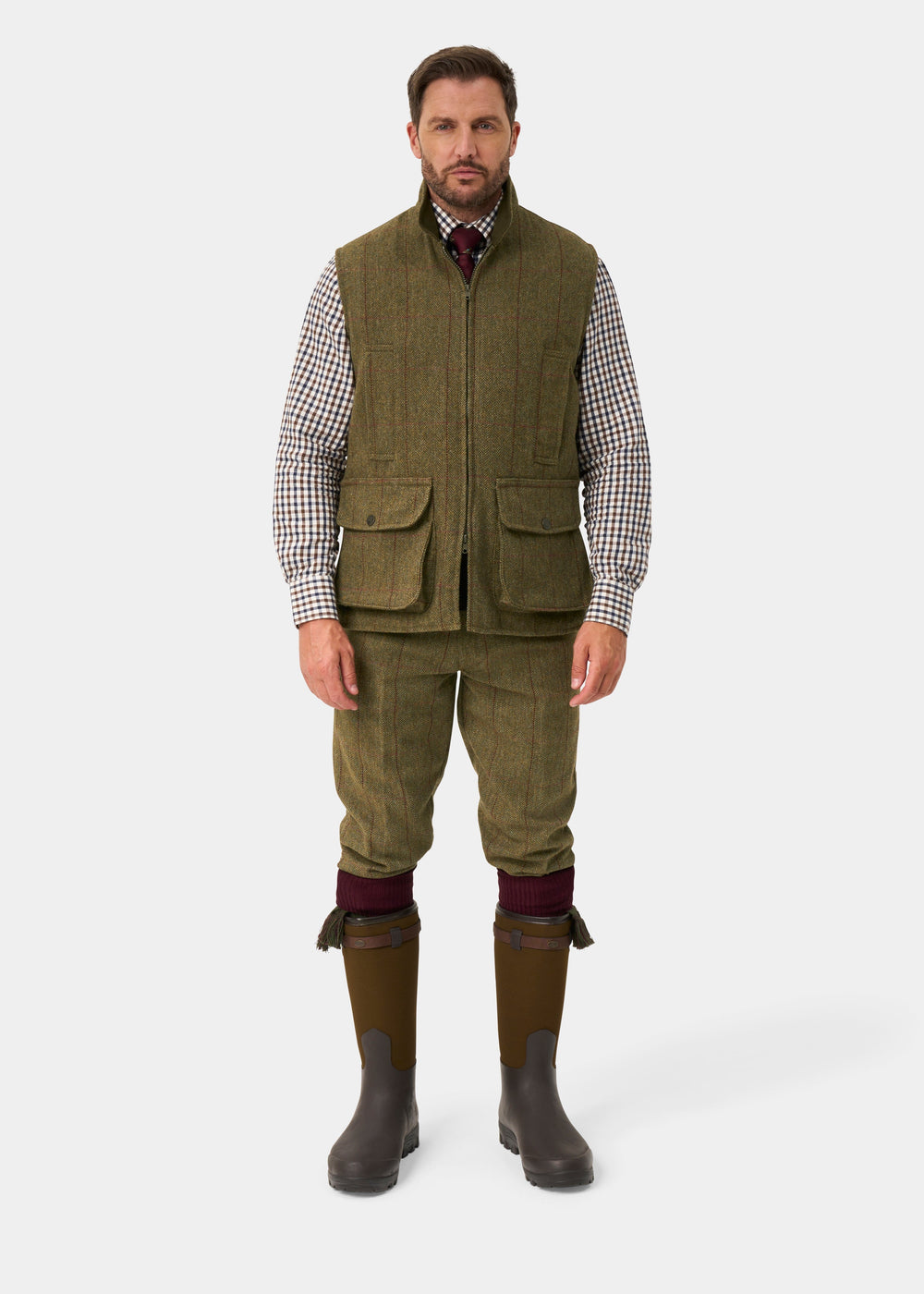 A man wears a Rutland Men’s Tweed Water Repellent Waistcoat In Oakley, matching breeks, checkered shirt, boots, and gaiters.