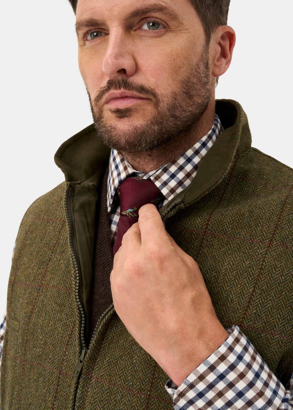 A man wears a Rutland Men’s Tweed Water Repellent Waistcoat In Oakley over a plaid shirt and dark red tie with a small pin.