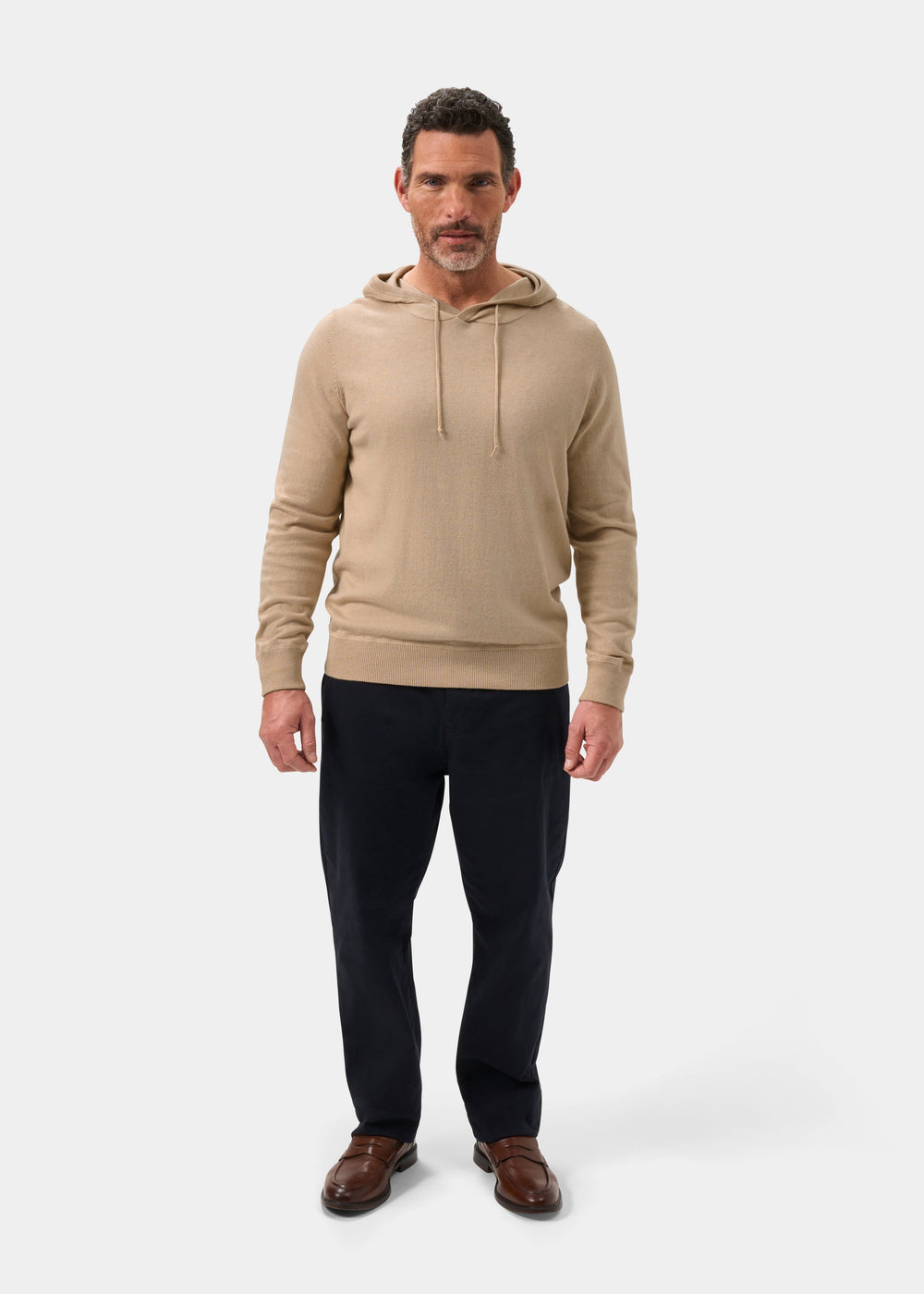 Man wears Gaisby Cotton Merino Hoodie in Oatmeal, dark pants, and brown shoes, standing forward against a plain background.