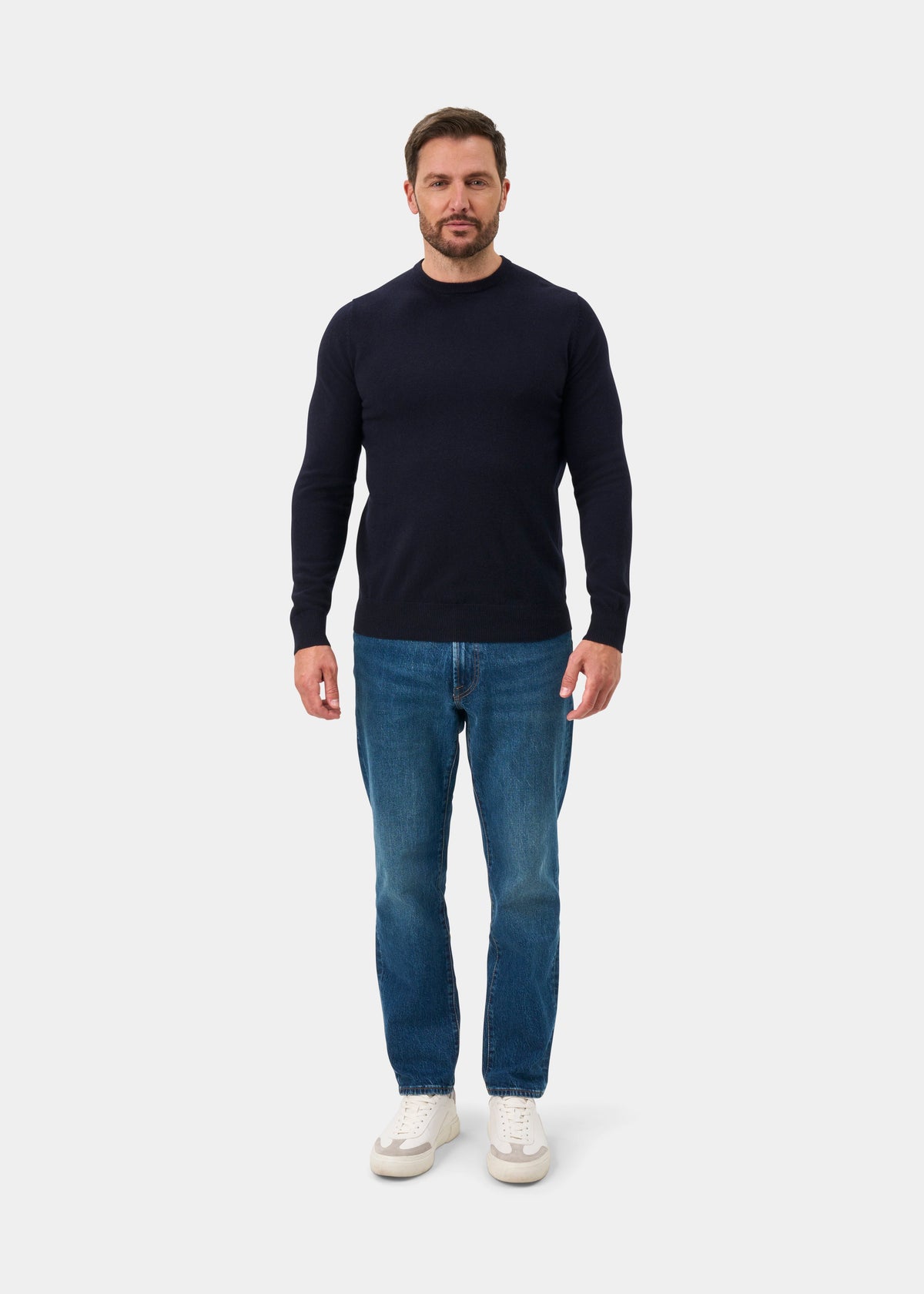 Man stands against a white background in a dark navy Brisbane Geelong Wool Crew Neck Jumper, blue jeans, and sneakers.