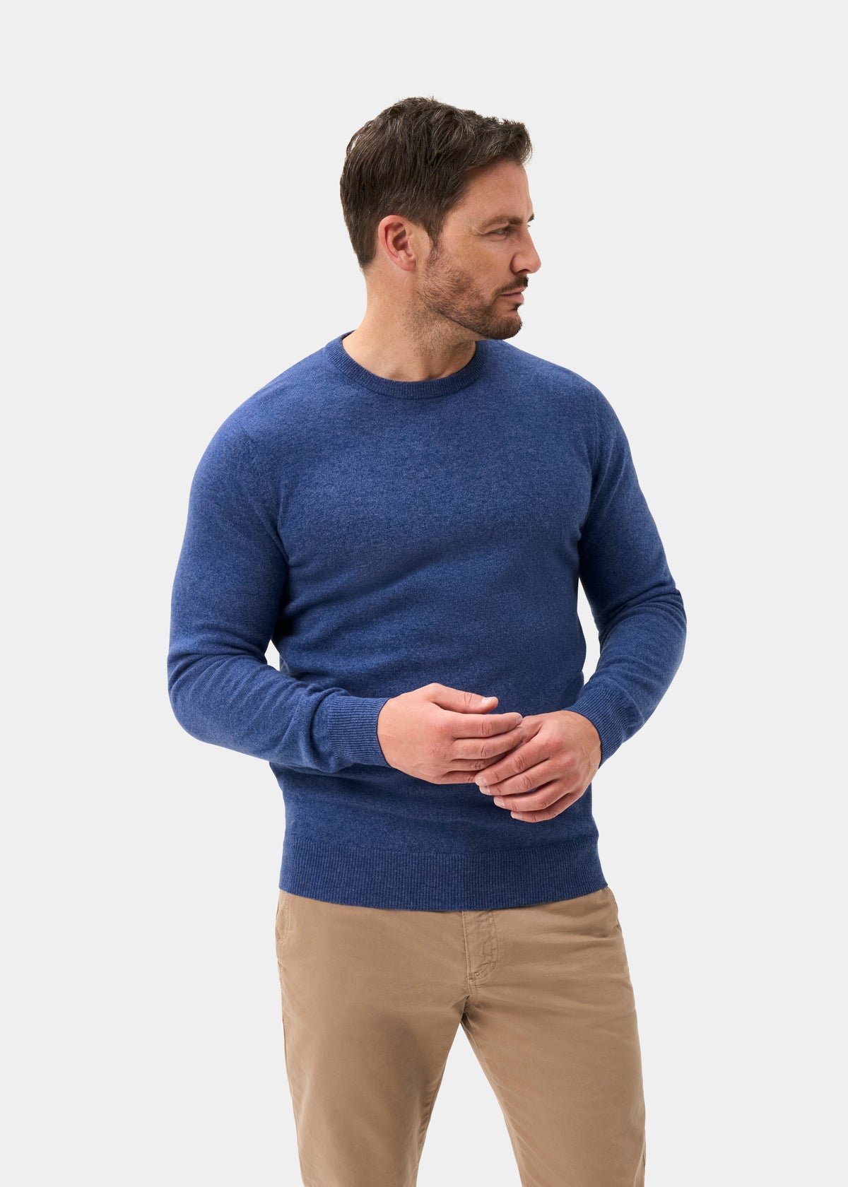Man in a blue Brisbane Geelong Wool Crew Neck Jumper and khaki pants stands, looking left against a plain background.