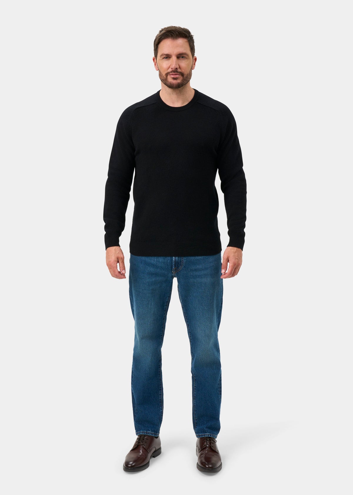 Man wears Lenzie Lambswool Crew Neck Jumper in black, blue jeans, and brown shoes, standing neutrally on white.
