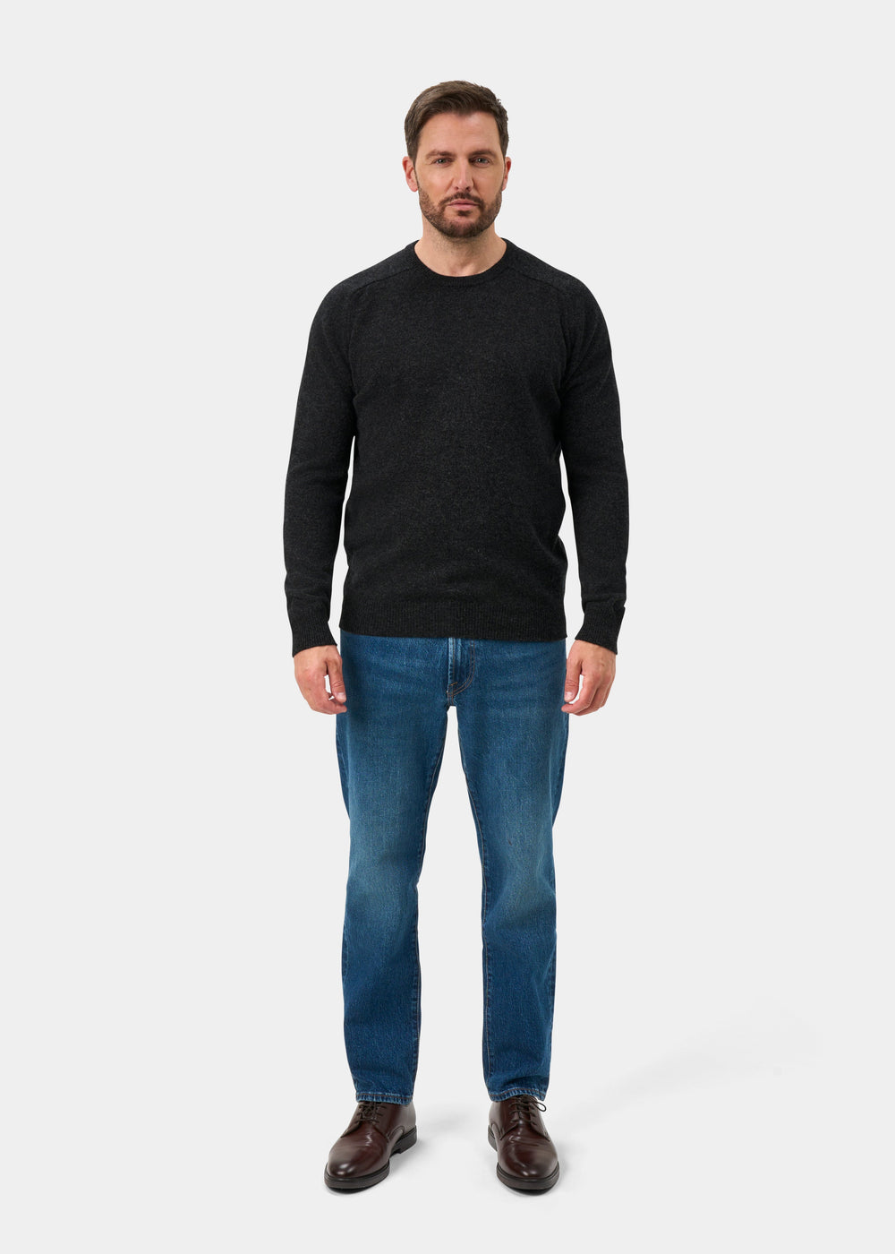 Bearded man wears Lenzie Lambswool Crew Neck Jumper in Charcoal, blue jeans, and brown shoes on white background.