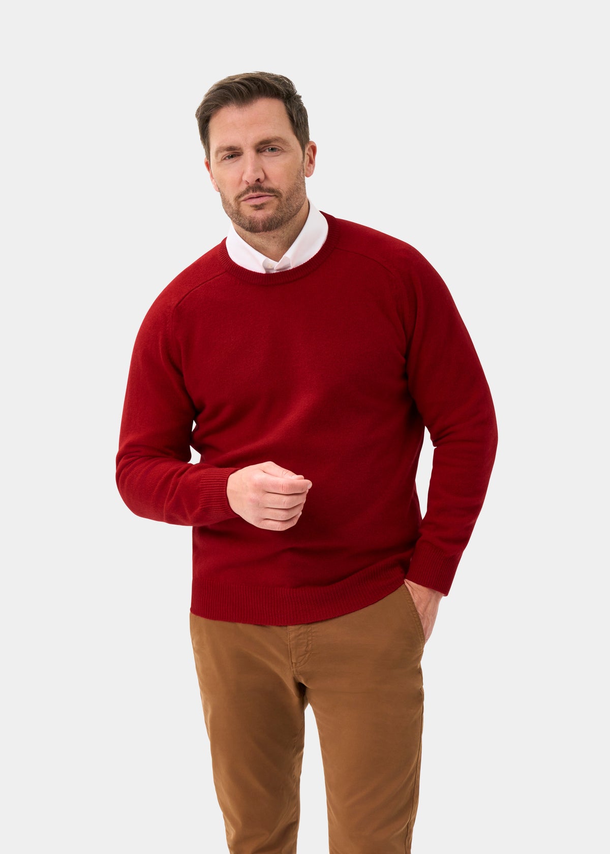 Man wears Dorset Lambswool Crew Neck Jumper in Dubonnet, classic fit, with brown pants against a plain white background.