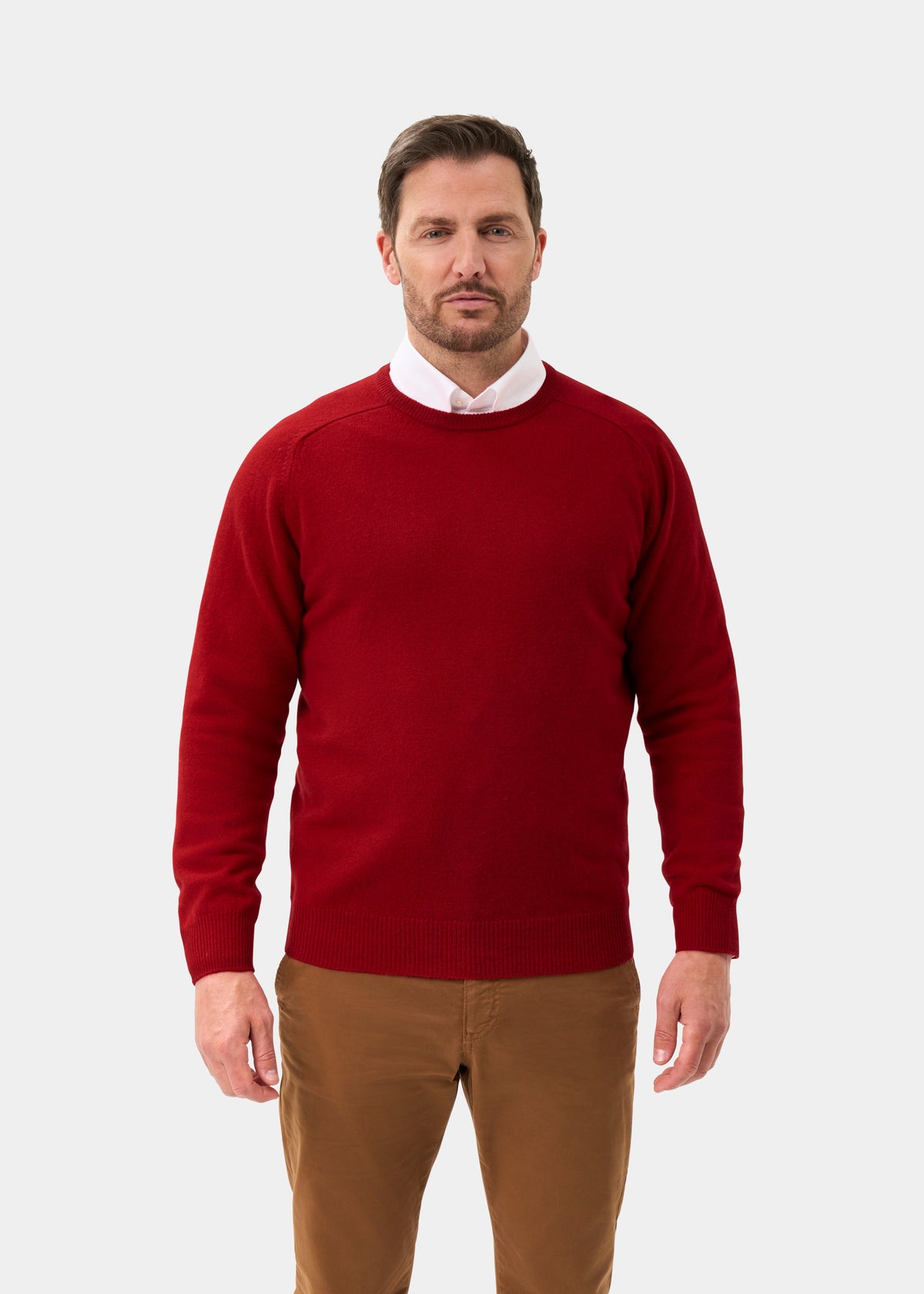 Man wears Dorset Lambswool Crew Neck Jumper in Dubonnet over white shirt, brown pants; stands against plain background.