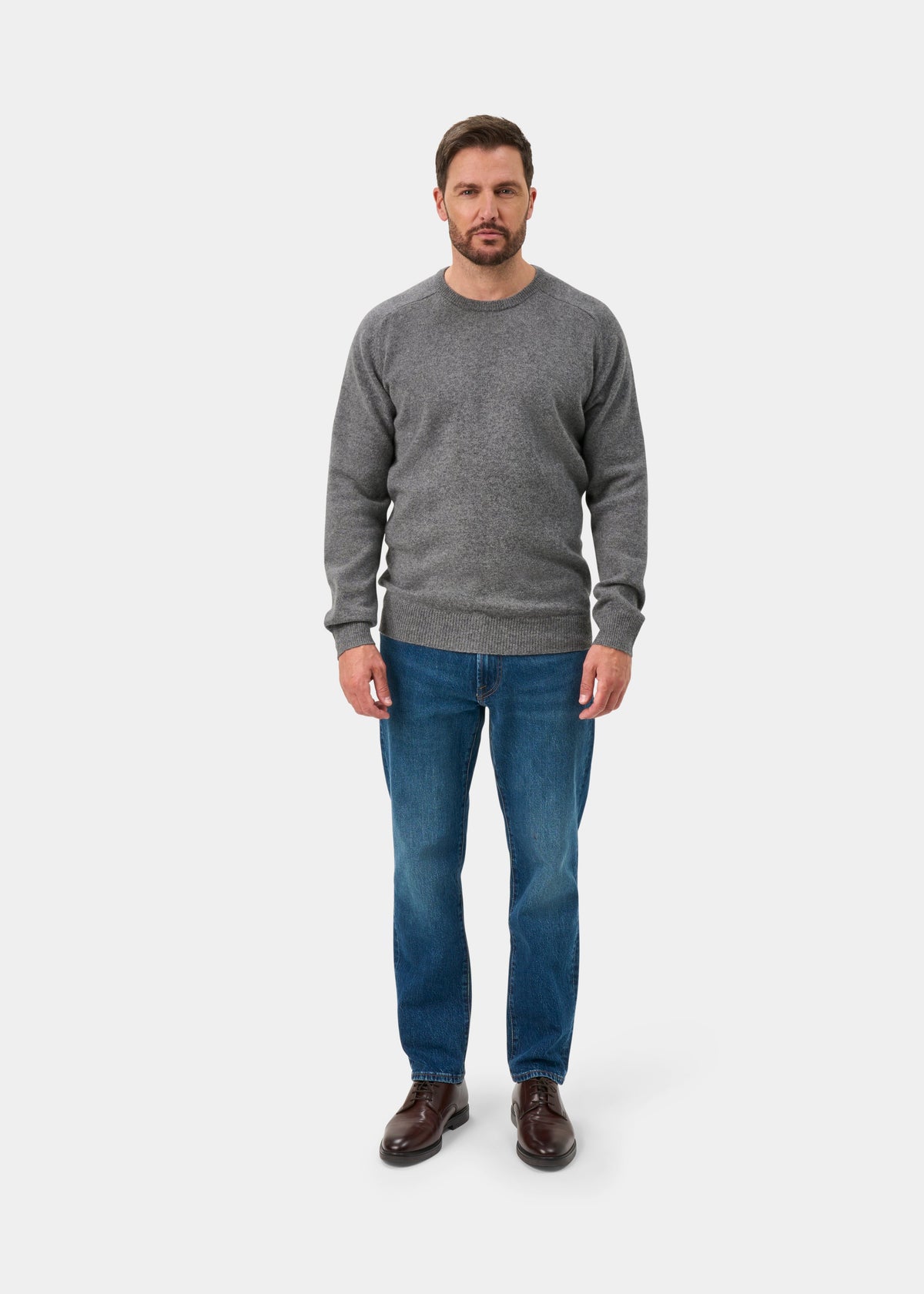 Man in Dorset Lambswool Crew Neck Jumper in Grey Mix, blue jeans, and brown shoes stands against a white background.