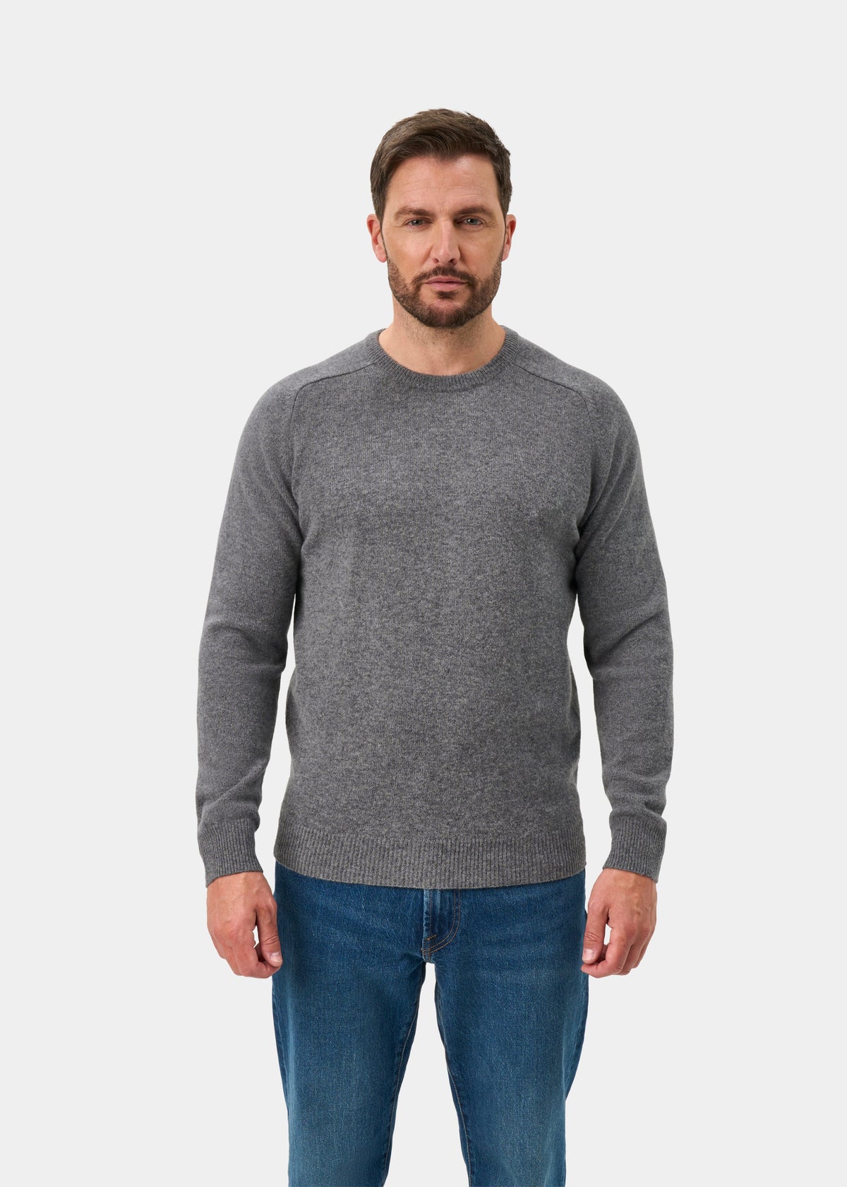 Man in Lenzie Lambswool Crew Neck Jumper (Grey Mix, Regular Fit) and blue jeans stands facing a white background.