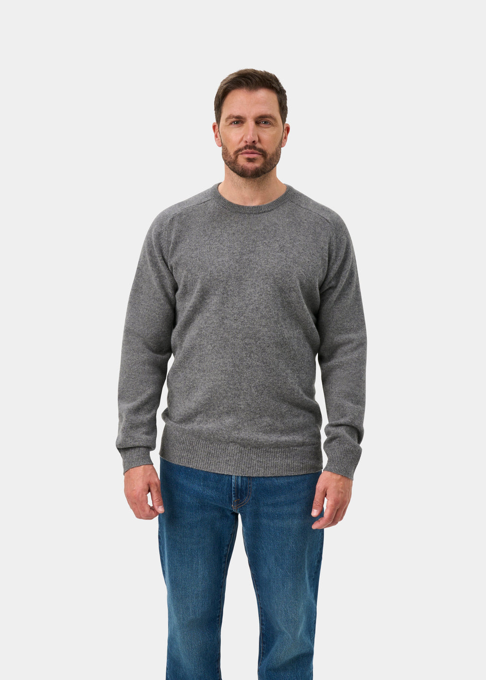Man wears Dorset Lambswool Crew Neck Jumper in Grey Mix, classic fit, with blue jeans, facing forward on white background.