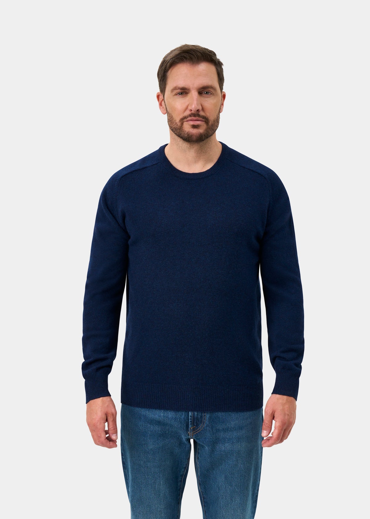 Man wears the Dorset Lambswool Crew Neck Jumper in Indigo with blue jeans against a plain light background.