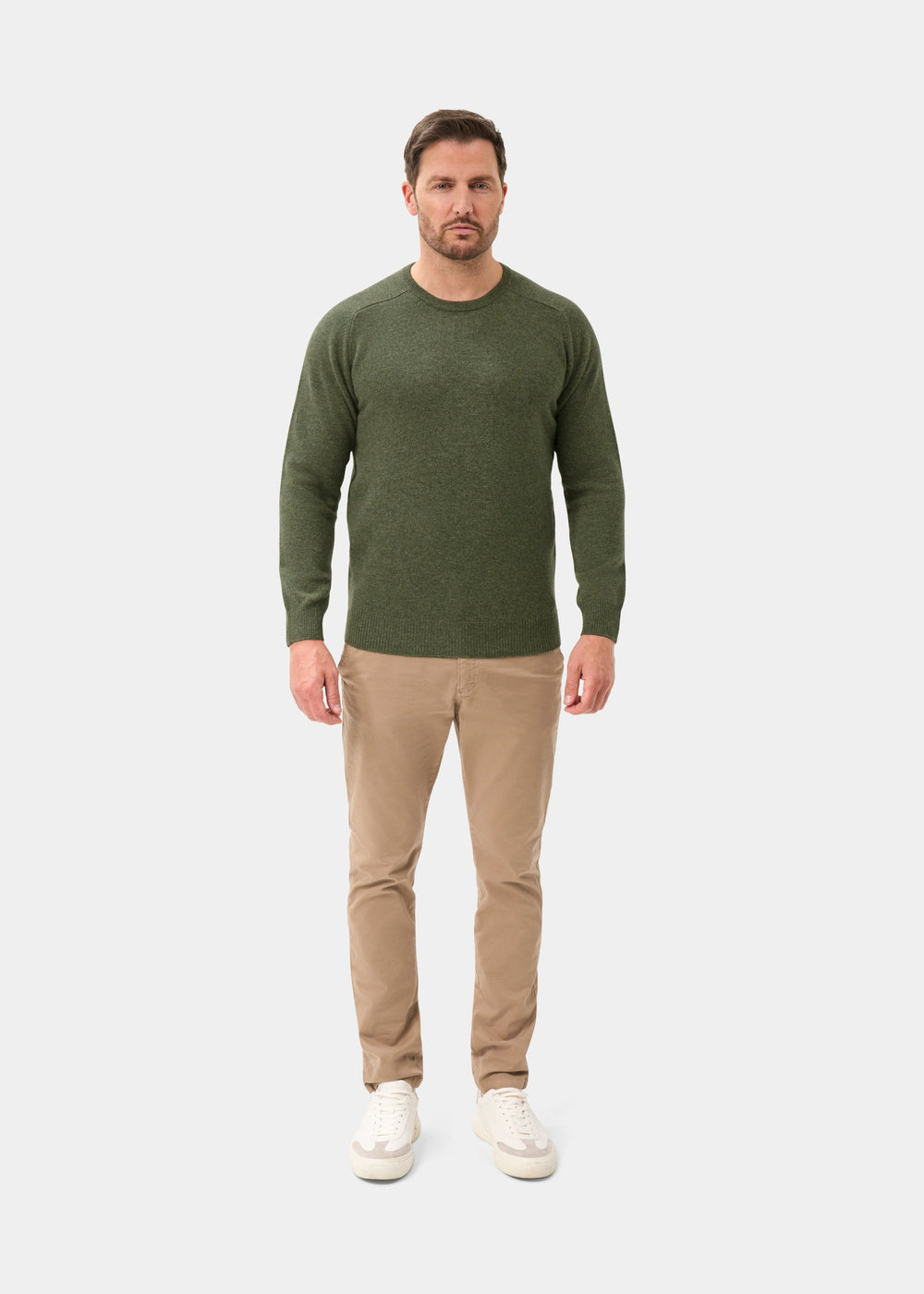 Man in green Lenzie Lambswool Crew Neck Jumper, tan pants, and white sneakers on a plain white background.