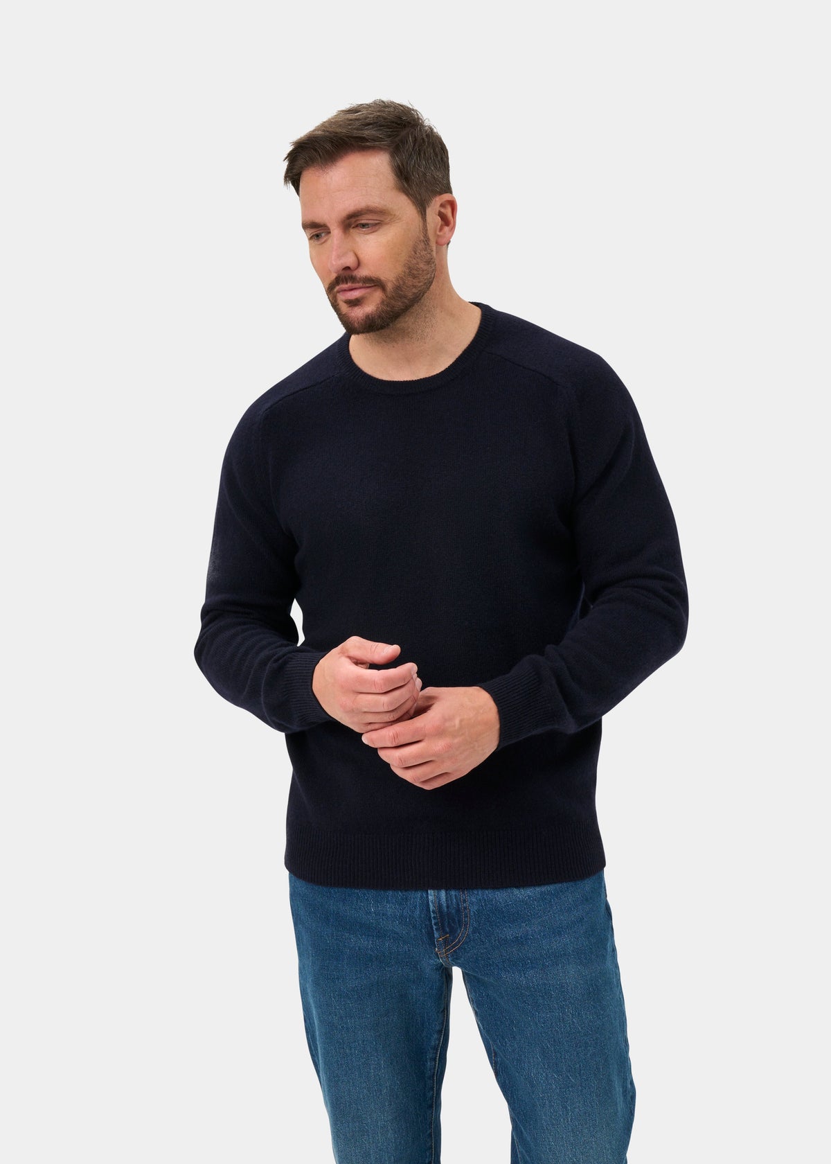 Man wears the Lenzie Lambswool Crew Neck Jumper in navy and blue jeans, standing against a plain light background.