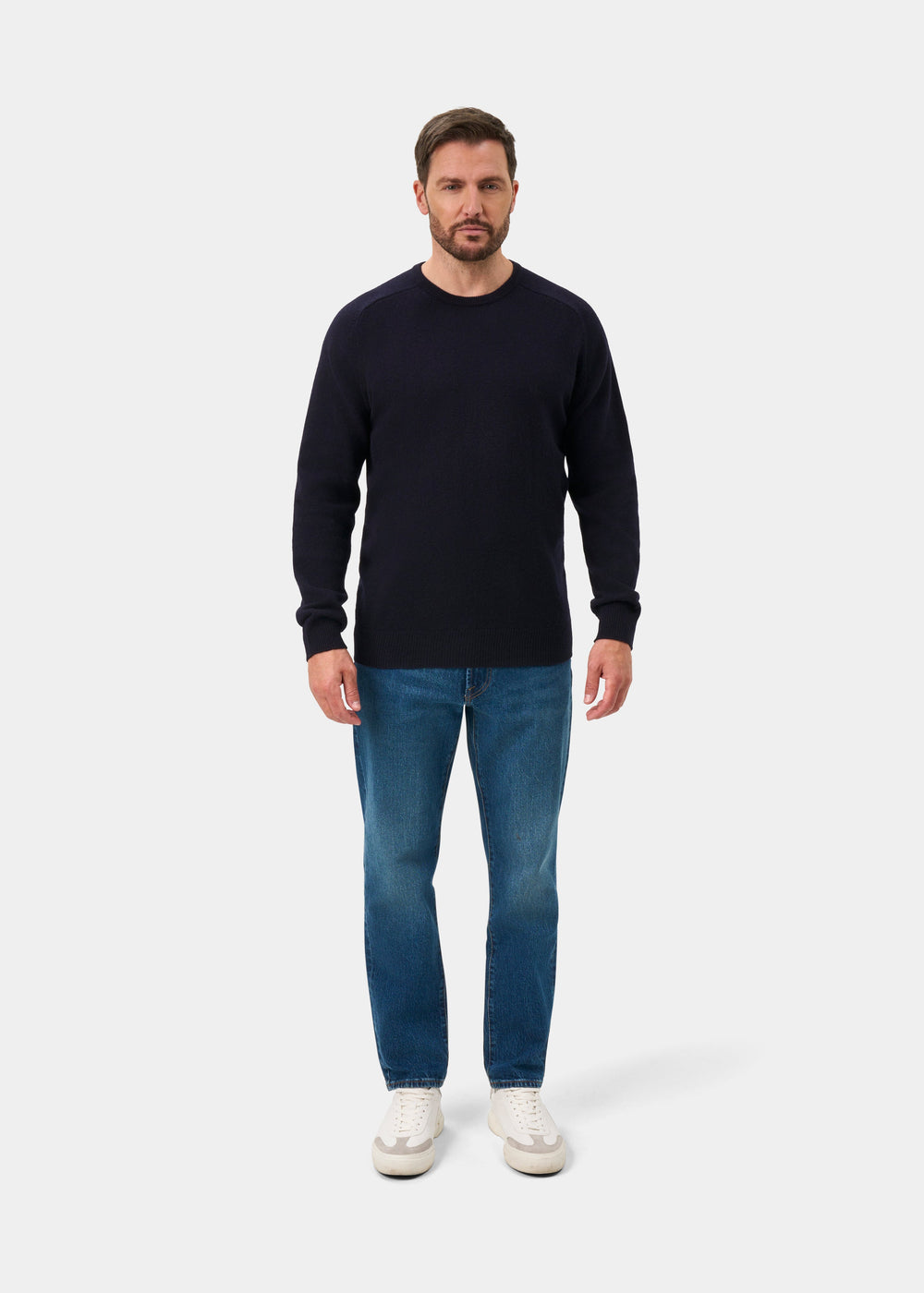 Man facing forward in Dorset Lambswool Crew Neck Jumper in navy, blue jeans, and white sneakers on white background.