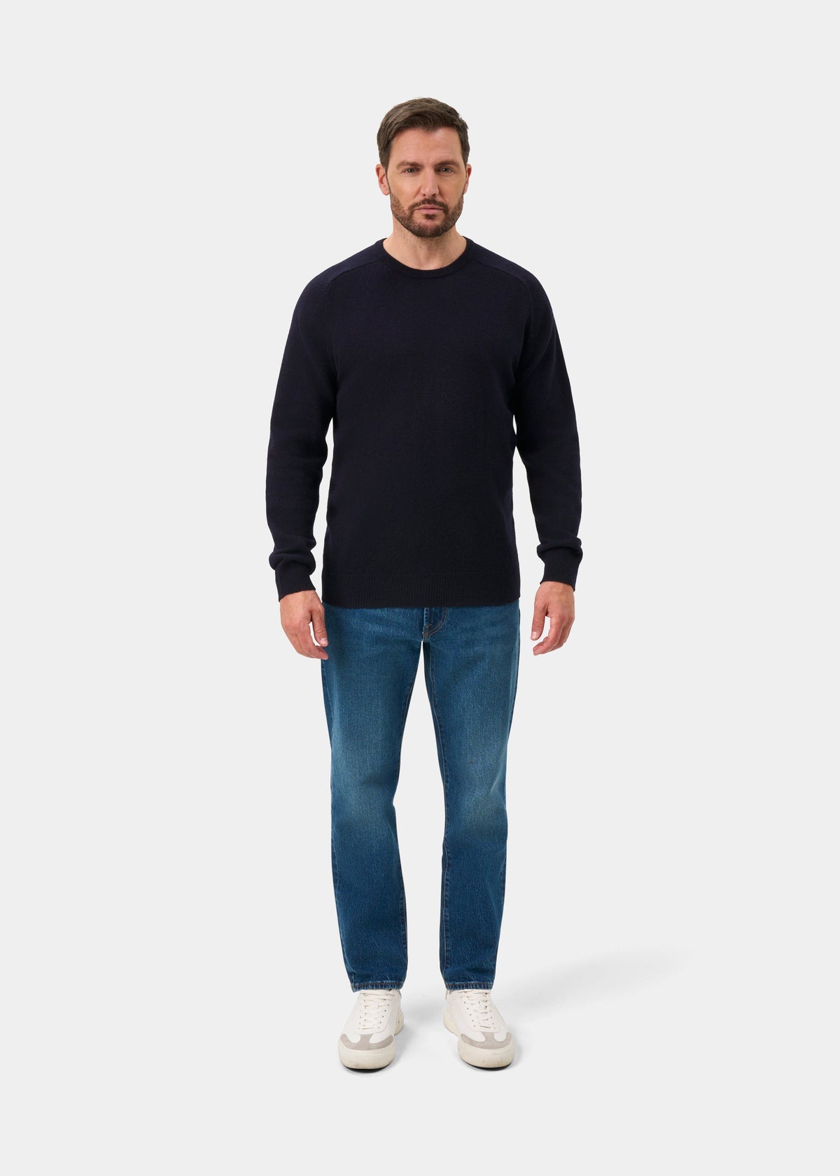 Man facing forward in Dorset Lambswool Crew Neck Jumper in navy, blue jeans, and white sneakers on white background.