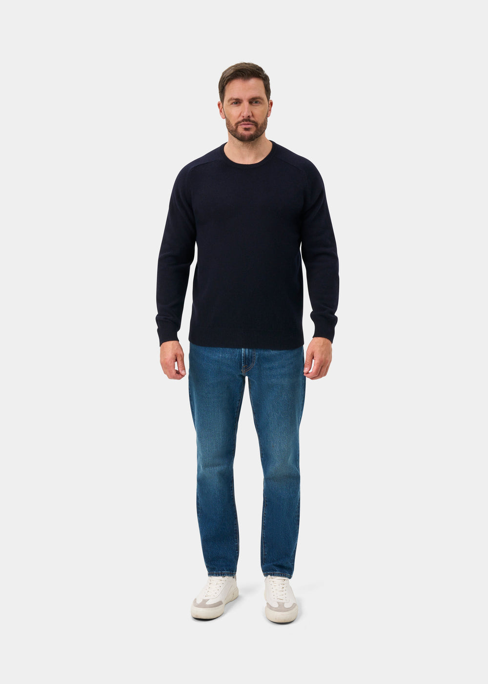 Man in Lenzie Lambswool Crew Neck Jumper in navy, blue jeans, and white sneakers, facing forward against plain backdrop.