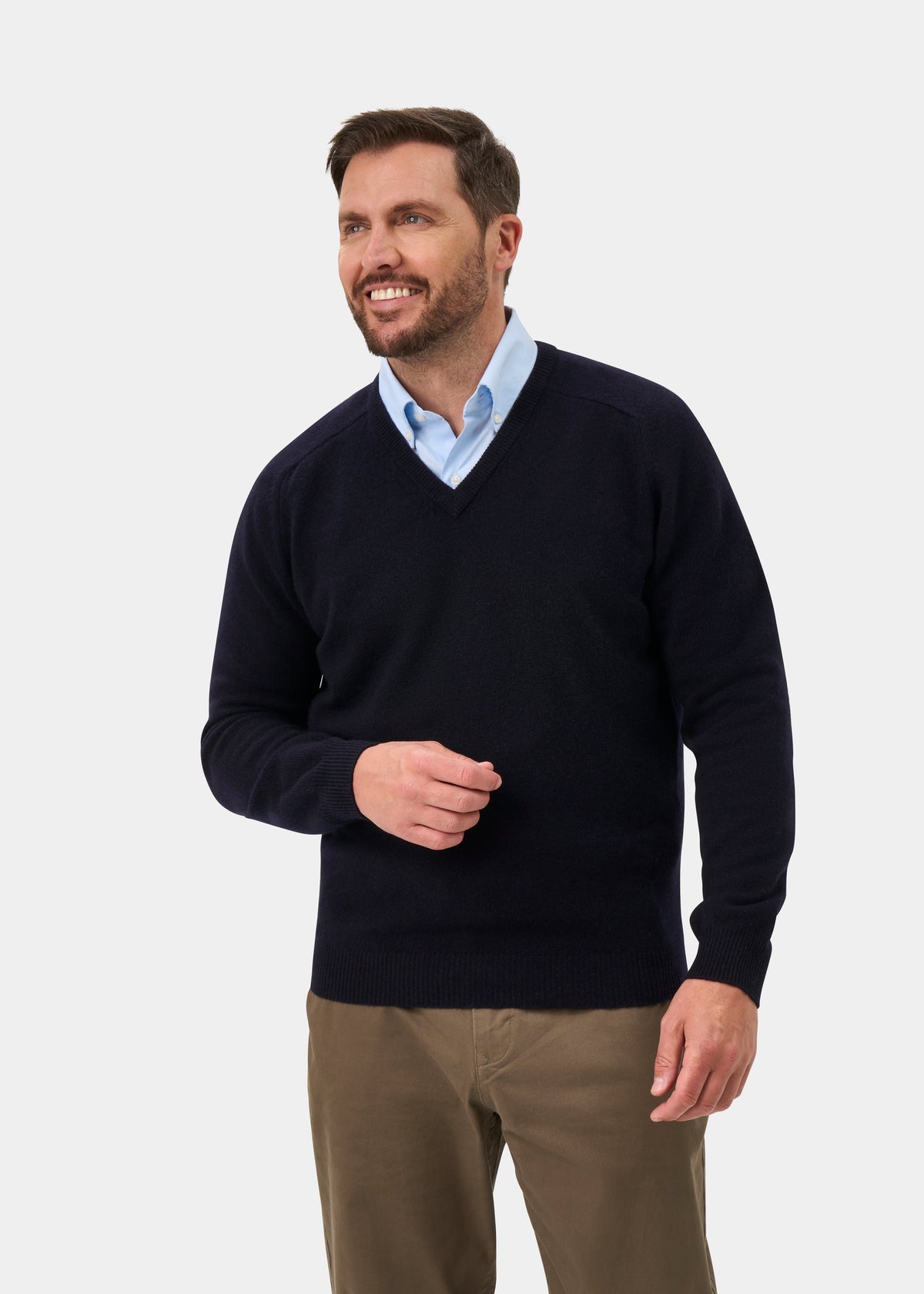 Man in Kilsyth Lambswool V-Neck Jumper in Navy, light blue shirt, khaki pants, smiles against plain backdrop.