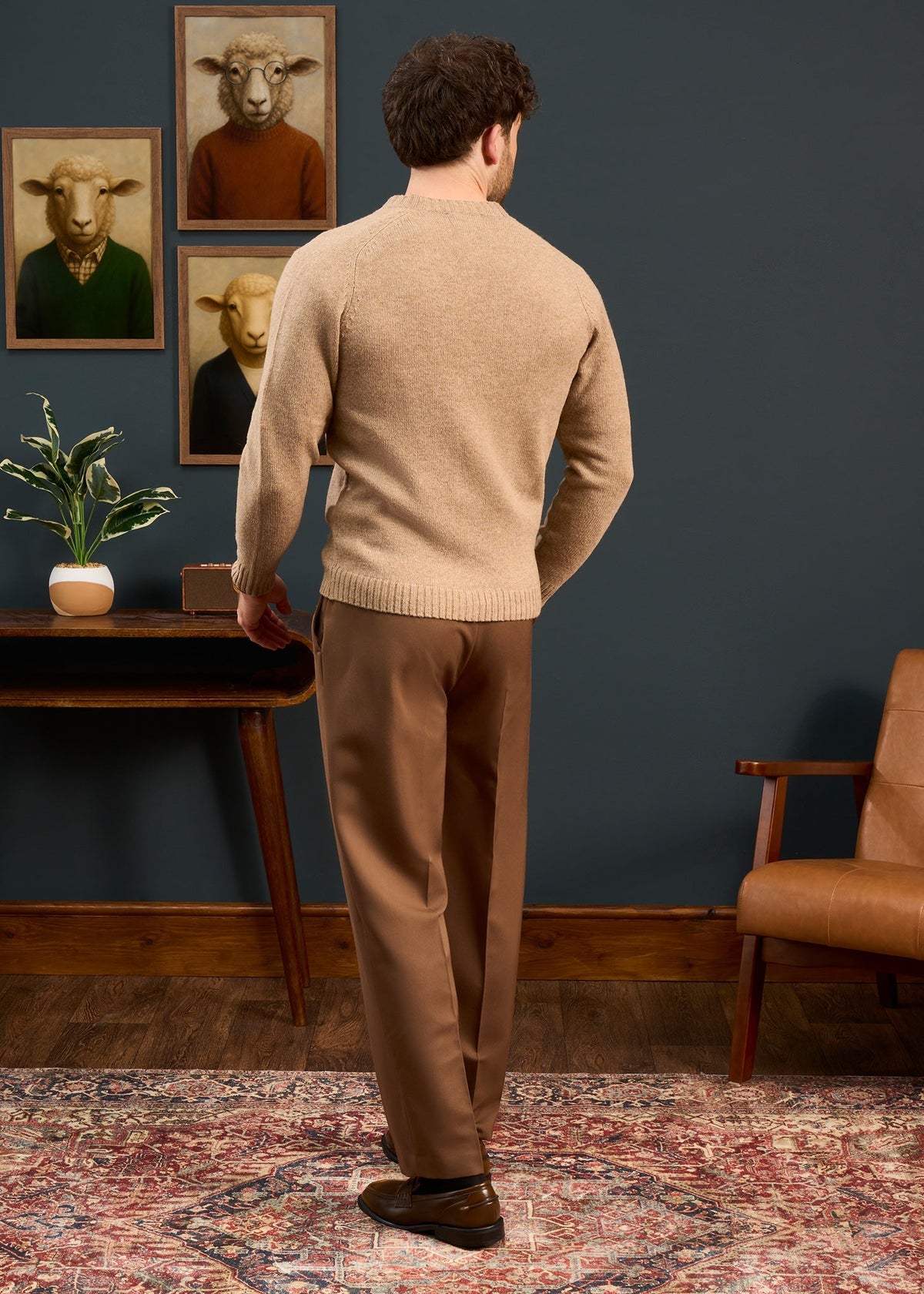 Man in Kinloch Supersoft Shetland Wool V-Neck Cardigan, pale oak, and brown pants stands with animal art, plant, and rug.