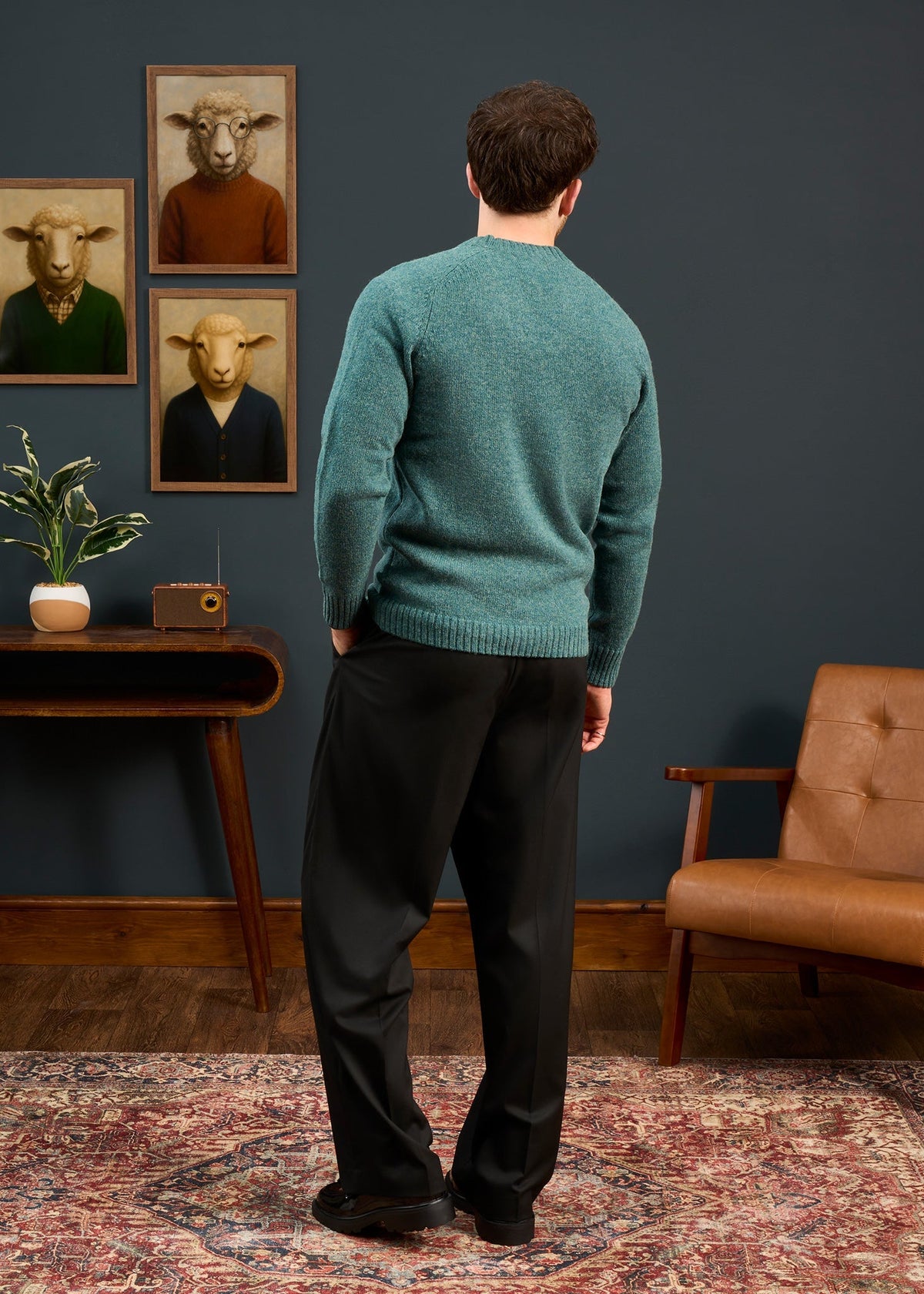 Man in a Kinloch Supersoft Shetland Wool V-Neck Cardigan in Kingfisher stands by a table, facing sheep portraits.