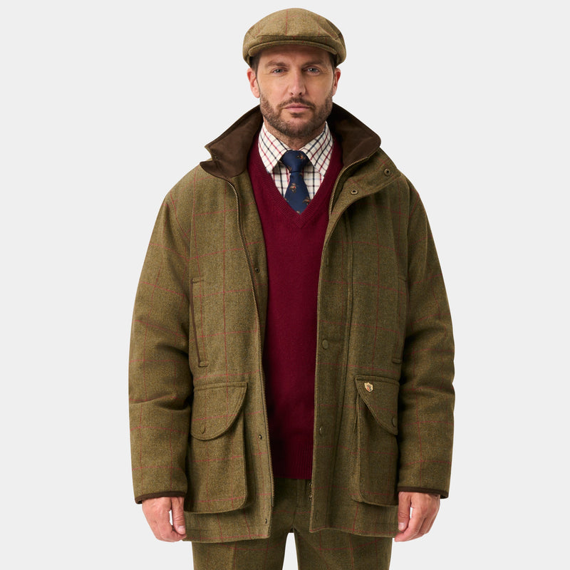 A man stands against a light gray studio background wearing an olive green hunting jacket, matching trousers, dark green rain boots, a burgundy sweater, and a brown flat cap.