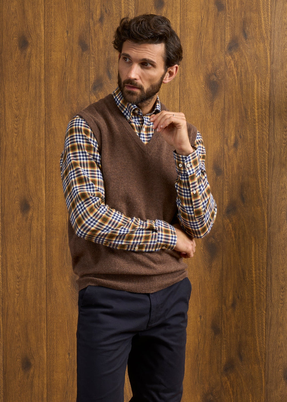 A man wears a Norfolk Lambswool V-Neck Slipover In Tobacco over a plaid button-down shirt.