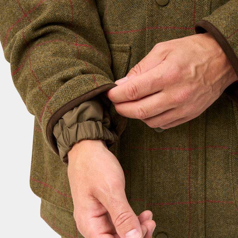 Close-up of a person wearing an olive green tweed jacket with a subtle red windowpane pattern, showing the elasticated cuff and hands adjusting it.
