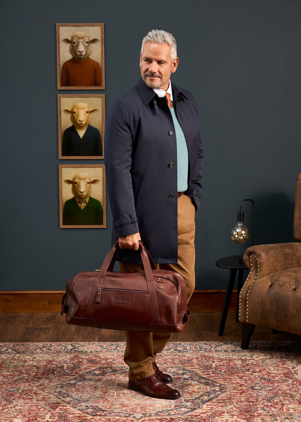 Alan Paine navy raincoat worn by a man holding a brown leather duffel bag in a sophisticated study setting.