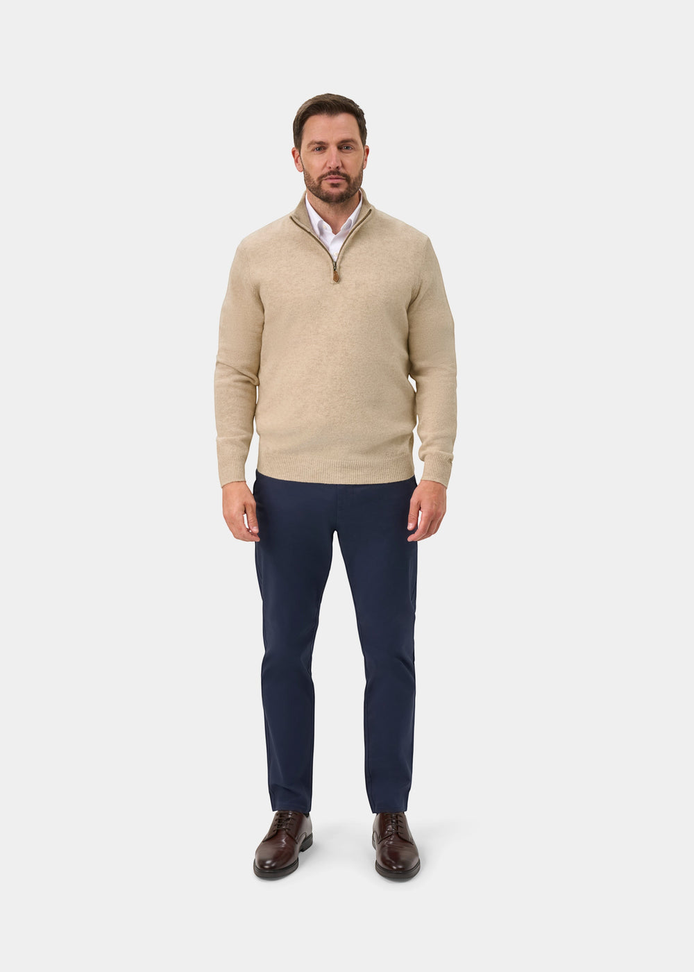 A man wears a Sutherland Lambswool Quarter Zip Jumper In Linen over a white collared shirt, navy trousers, and brown shoes.
