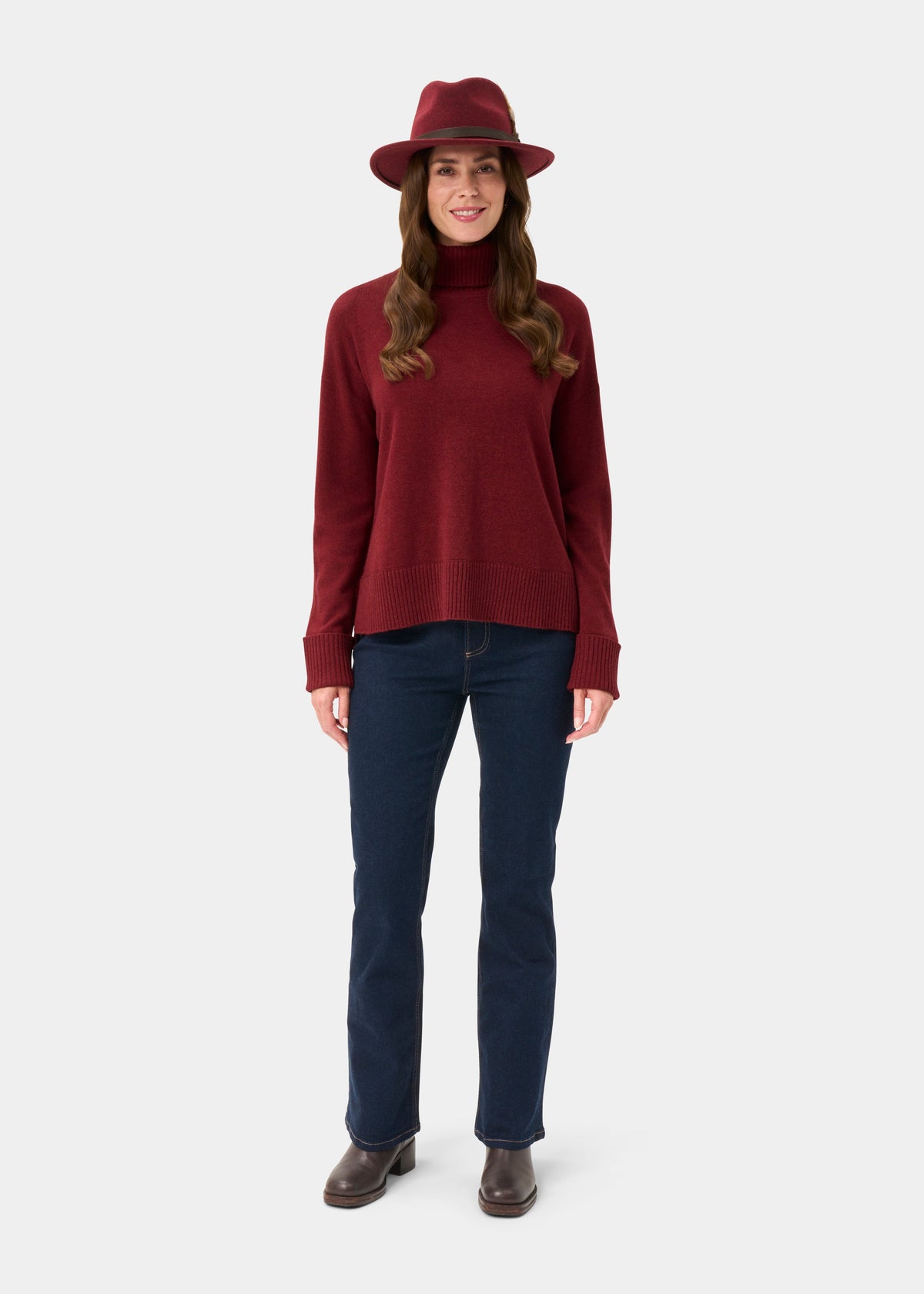Woman in Alexa Ladies Berry Roll Neck, red hat, blue jeans, and brown boots smiles against a white background.