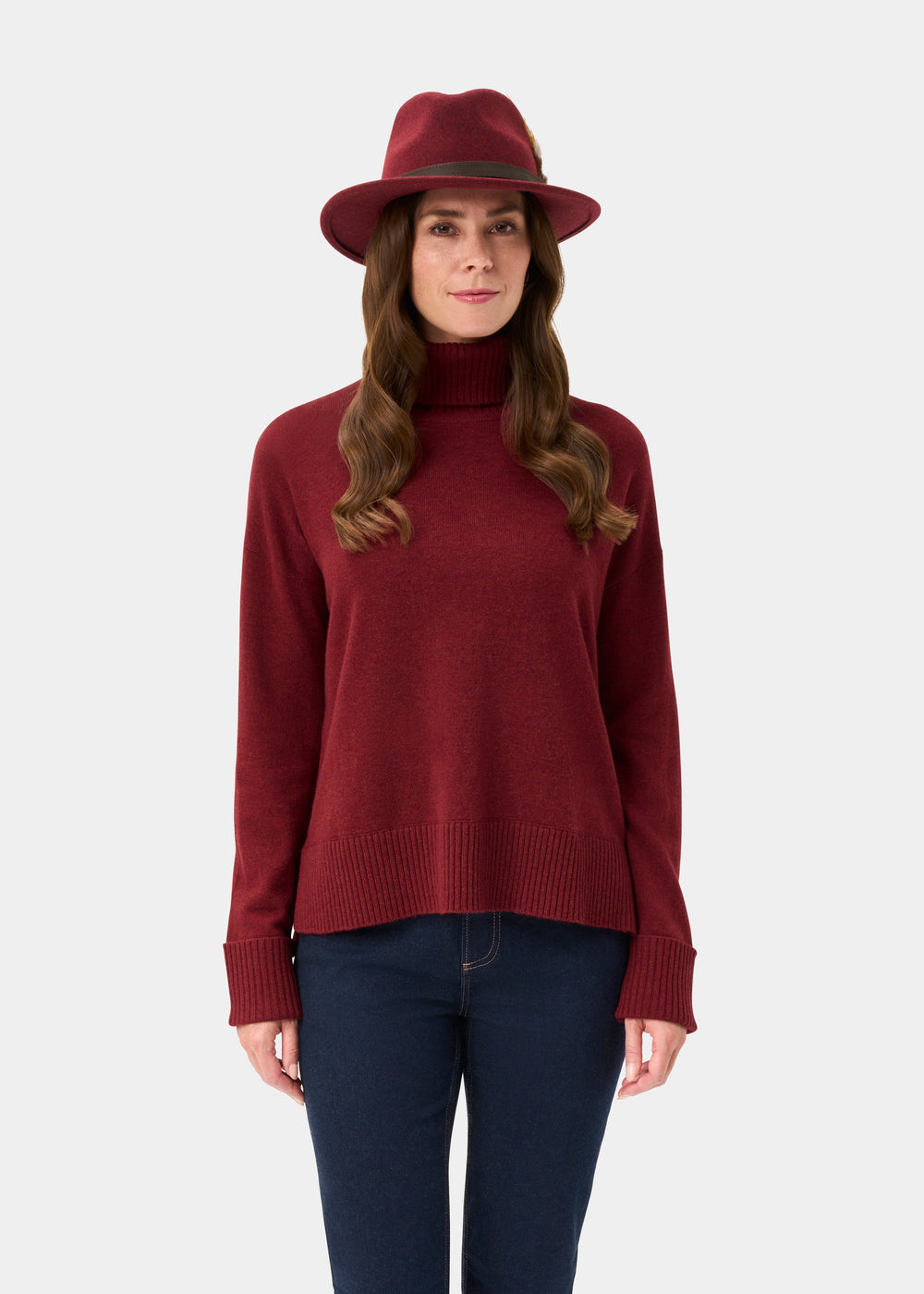 Woman in Alexa Ladies Wool and Cashmere Roll Neck in Berry, red hat, and blue jeans stands against a plain background.