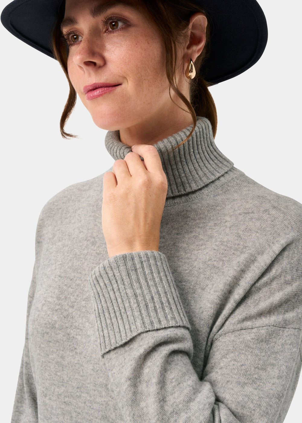 Woman in the Alexa Wool & Cashmere Roll Neck Jumper in Husky, black hat, touching collar, looking to the side.