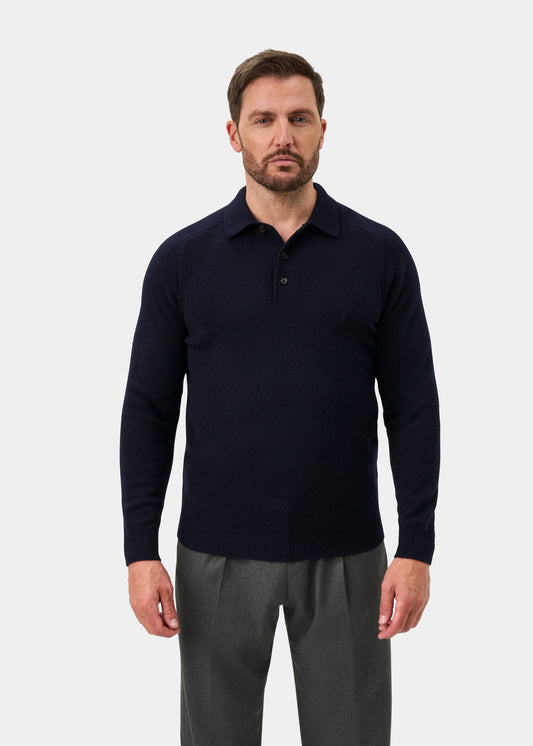 Man wears Bilsdean Men’s Lambswool Long Sleeve Polo in Navy, regular fit, with gray trousers against a plain backdrop.