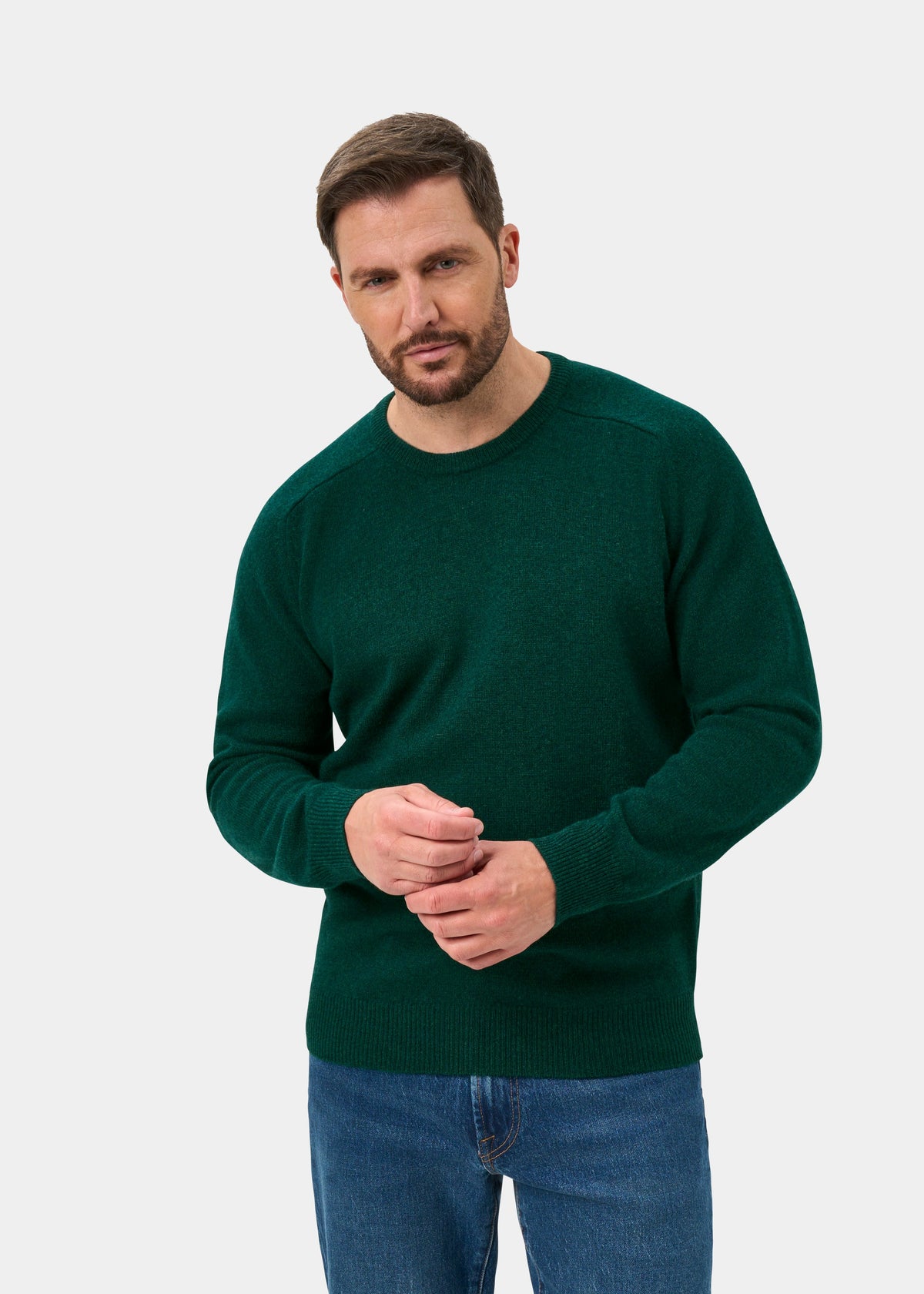 Man wears Dorset Lambswool Crew Neck Jumper in Mallard, classic fit, with blue jeans, against a white background.