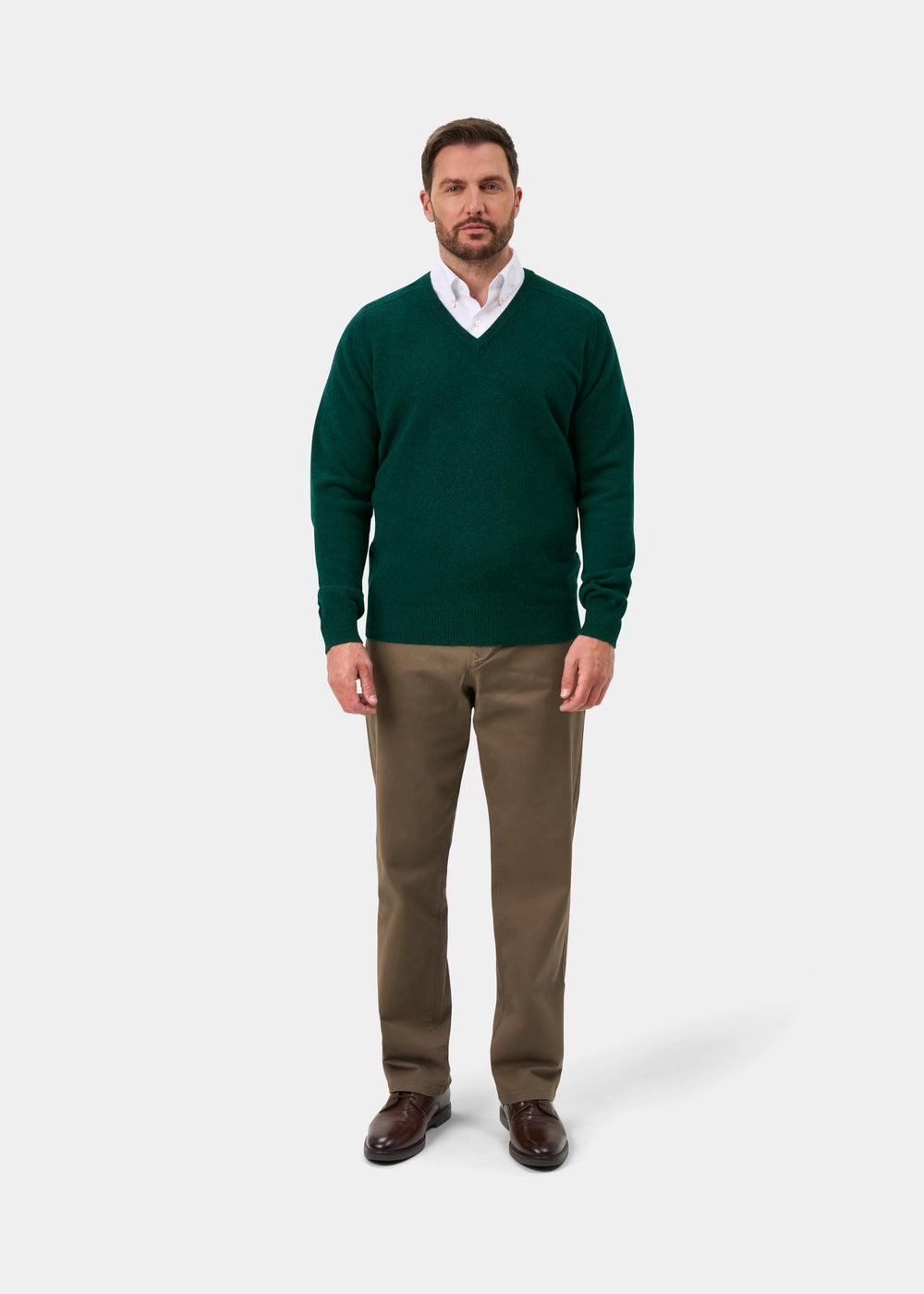 Man in Hampshire Lambswool V-Neck Jumper in Mallard, white shirt, brown pants & shoes stands by plain white background.
