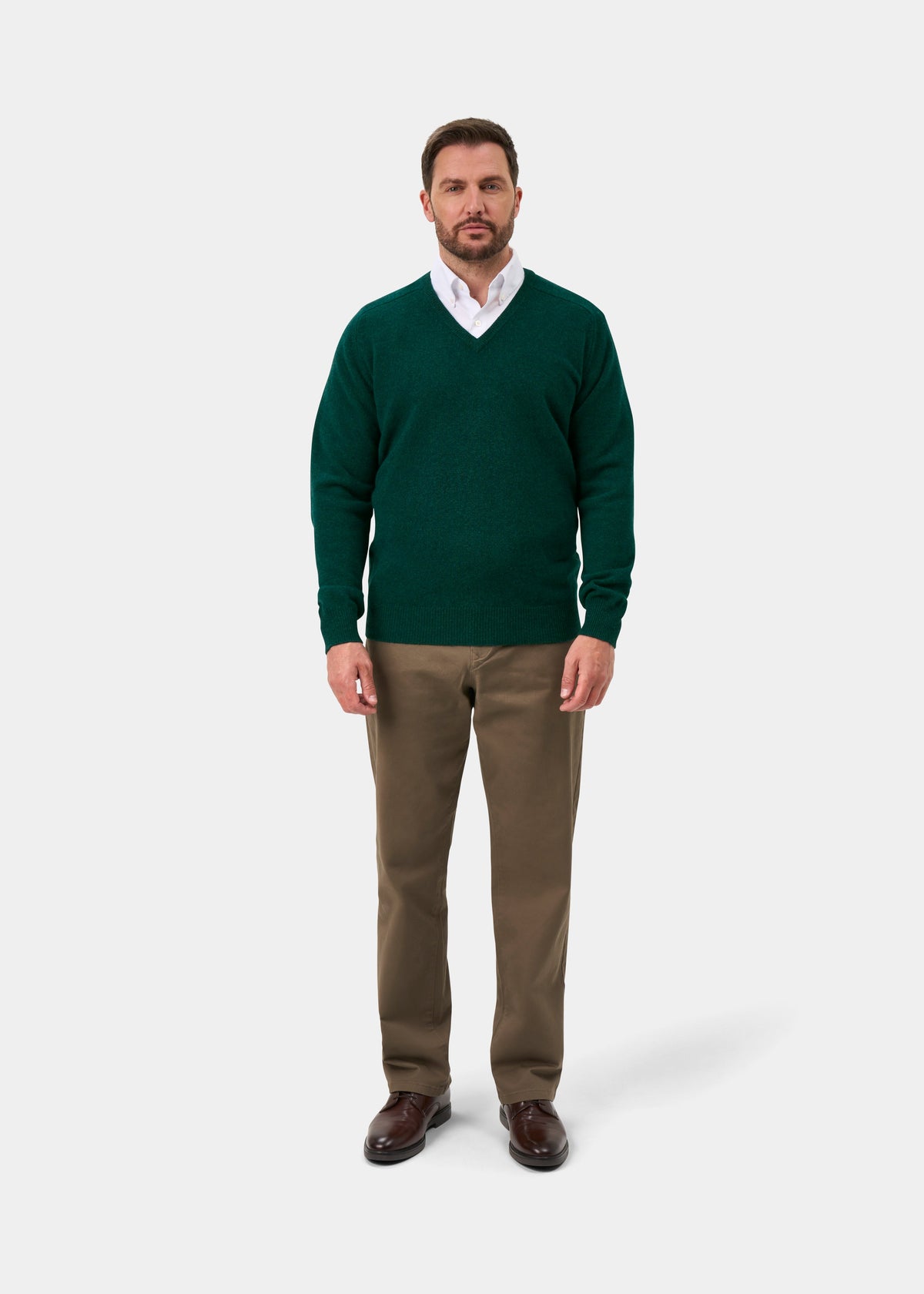 Man in Hampshire Lambswool V-Neck Jumper in Mallard, white shirt, brown pants & shoes stands by plain white background.