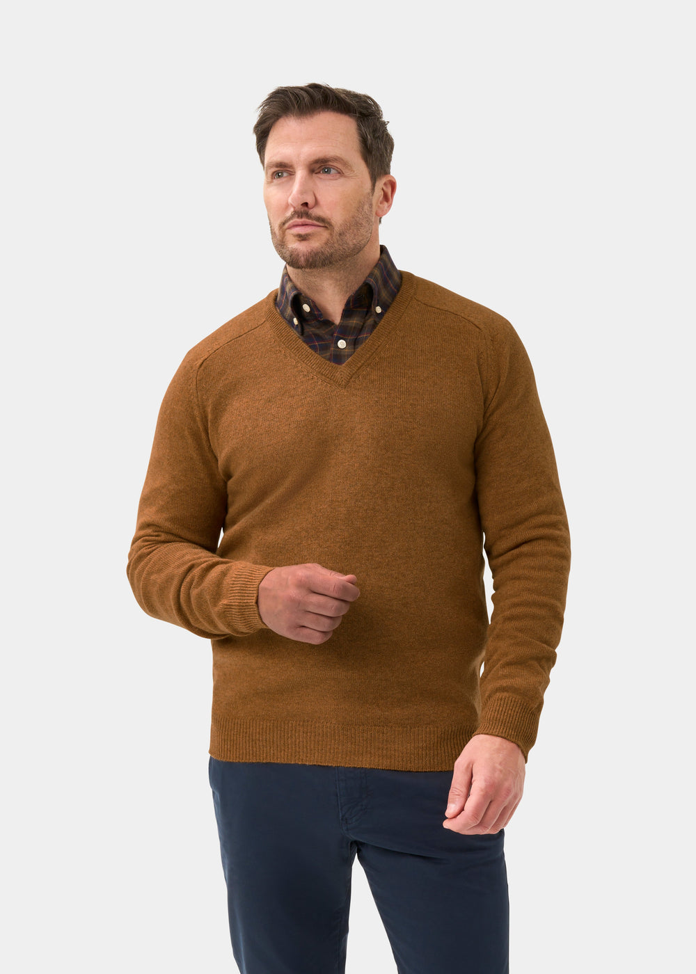 Man wears Kilsyth Lambswool V-Neck Jumper in Gazelle over polka dot shirt, navy pants; light backdrop.