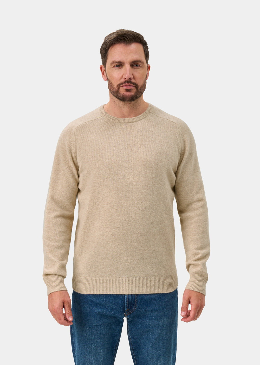 Man with short brown hair wears the Lenzie Lambswool Crew Neck Jumper in Linen, paired with blue jeans, on white.