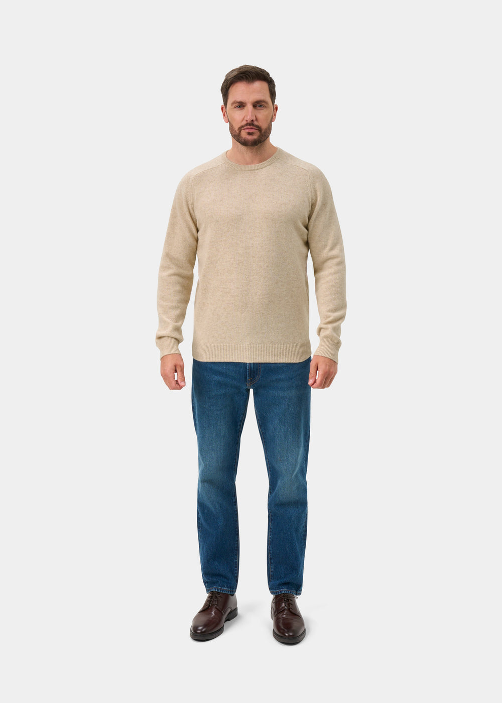 A man in a Lenzie Lambswool Crew Neck Jumper in Linen, blue jeans, and brown shoes stands facing forward.