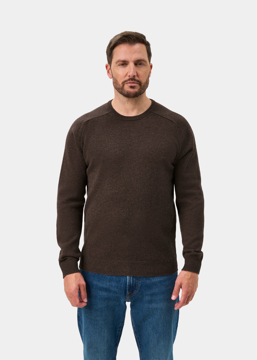 Man wears Lenzie Lambswool Crew Neck Jumper in mocha, regular fit, with blue jeans, facing forward on white background.