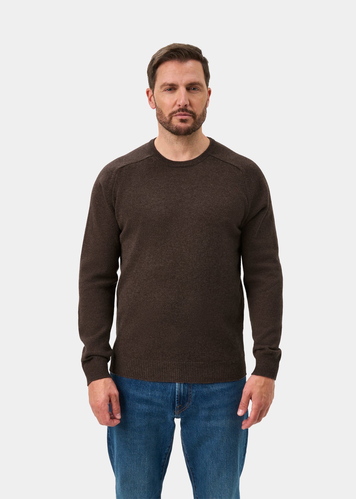 Man wears Lenzie Lambswool Crew Neck Jumper in mocha, regular fit, with blue jeans, facing forward on white background.