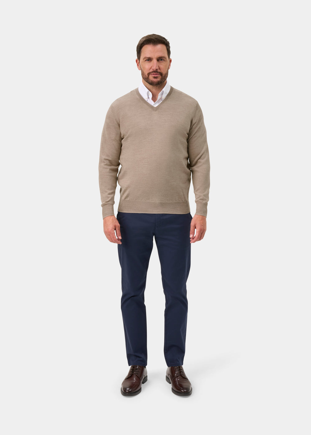 A man wearing a beige v-neck sweater over a white collared shirt, navy blue trousers, and brown leather shoes, standing against a light gray studio background.
