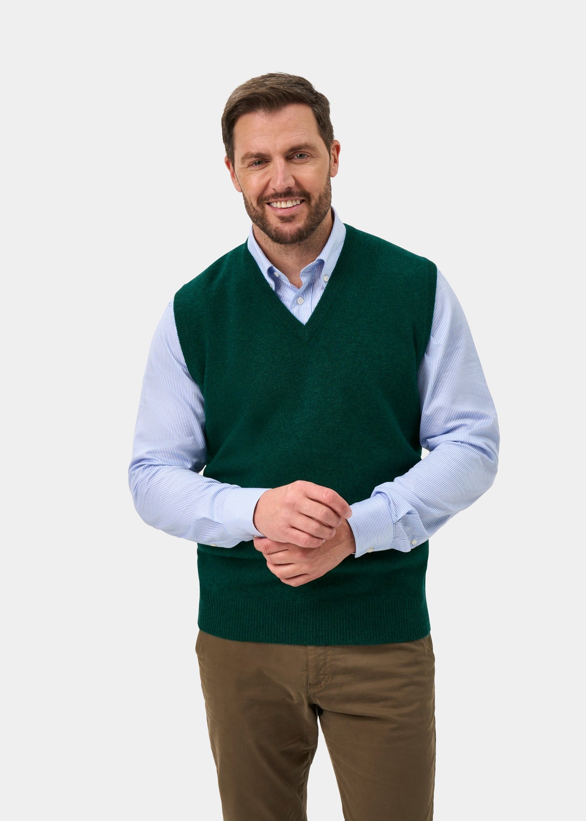 A smiling man wearing a dark green V-neck sweater vest over a light blue collared shirt and brown trousers, standing against a light gray studio background.