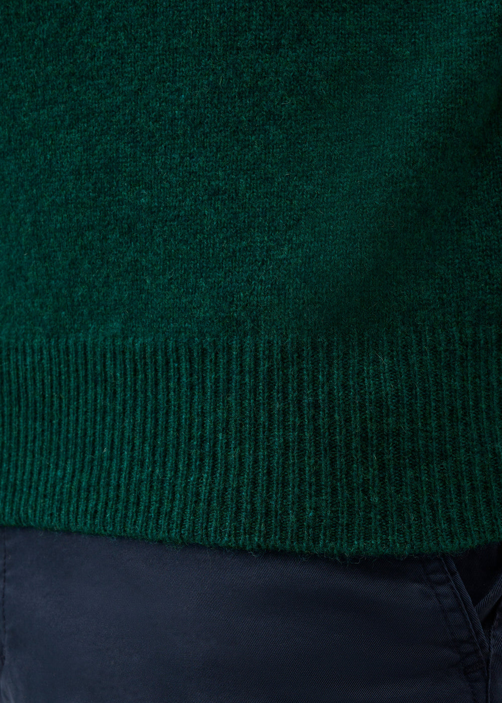 Close-up of a dark green knit sweater with a ribbed hem, worn over dark navy pants.