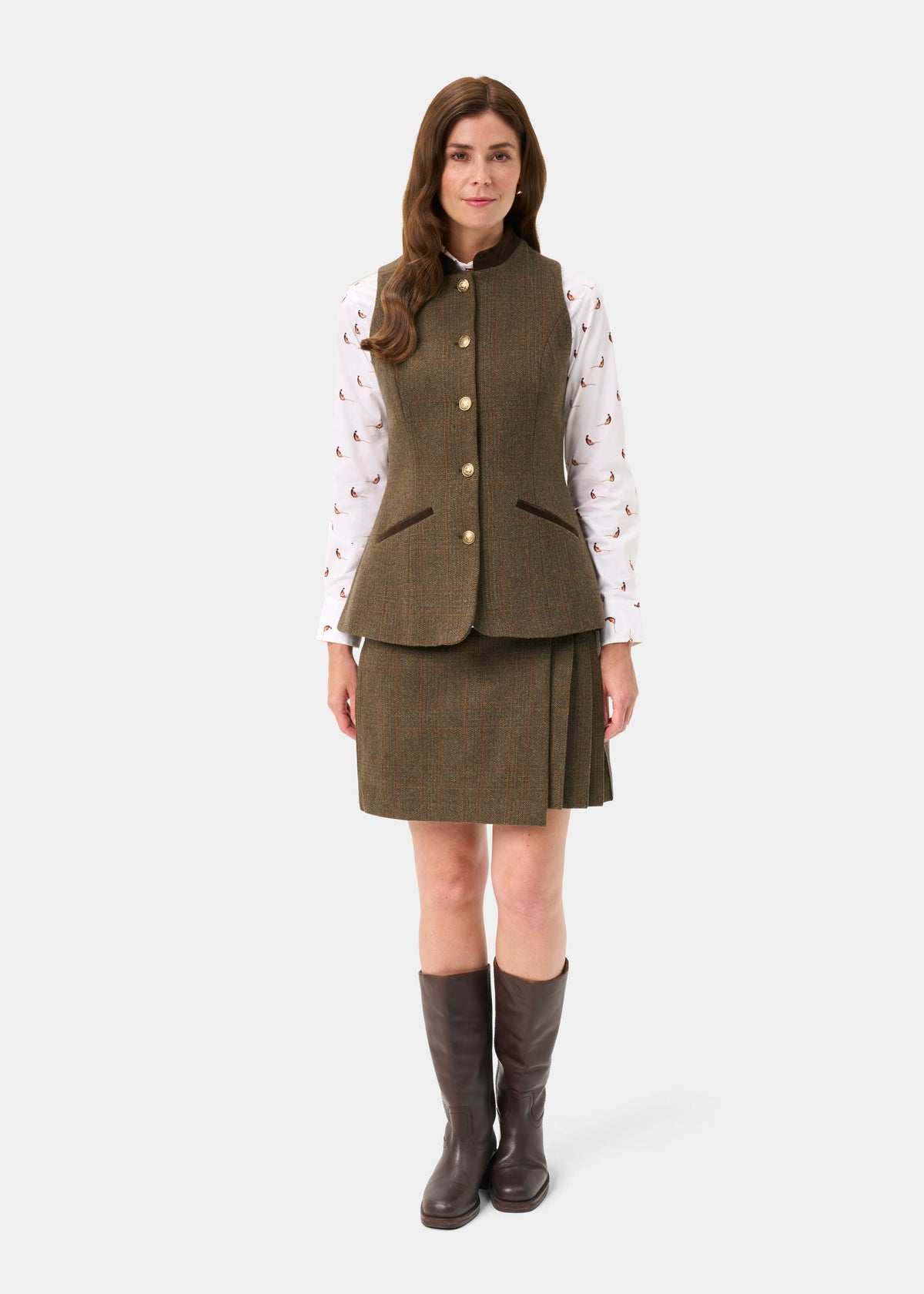 A woman stands against a light gray studio background wearing a brown tweed vest and matching pleated skirt over a white long-sleeve blouse with a small pink pattern, paired with dark brown knee-high boots.